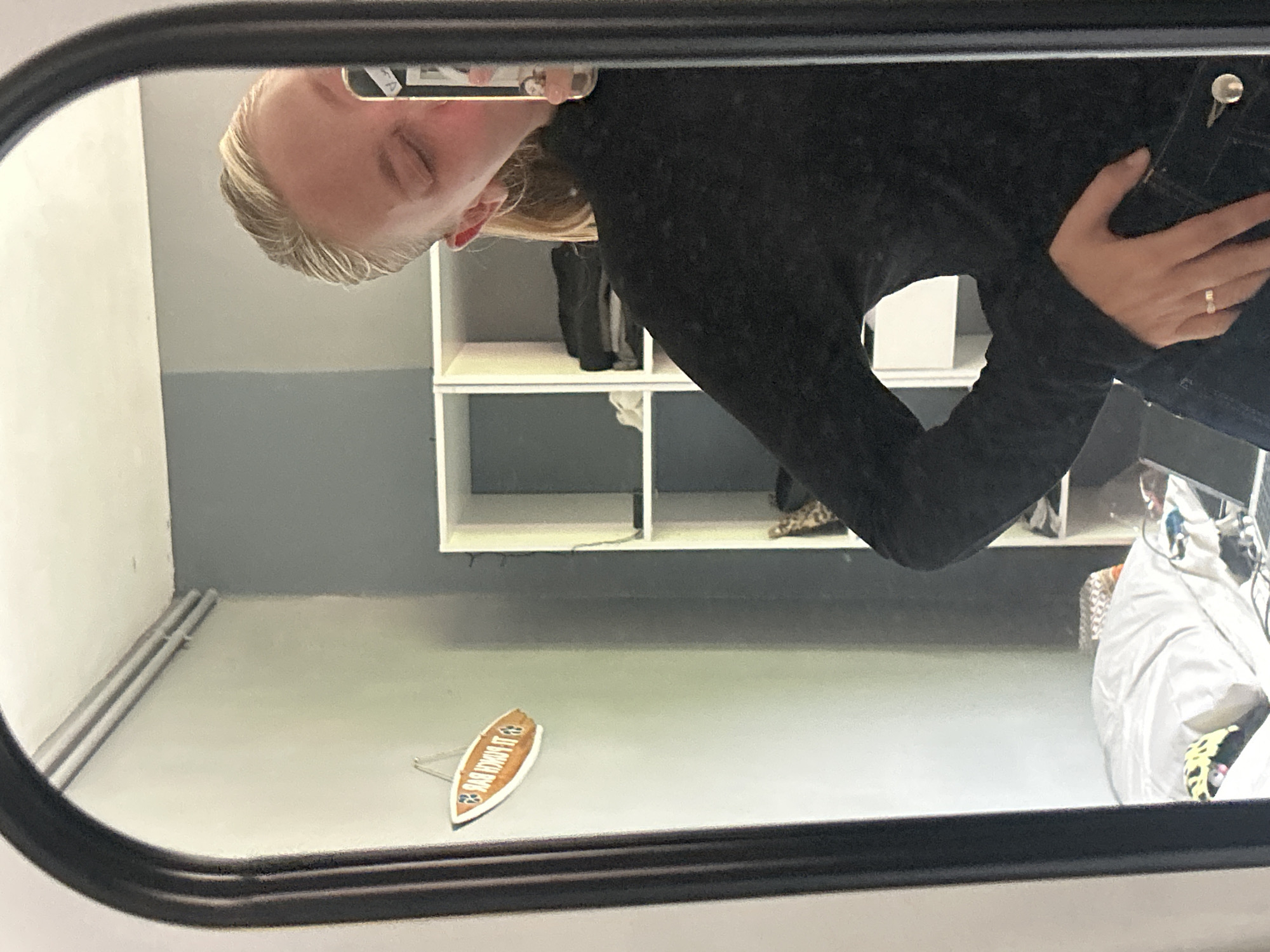 A mirror reflection shows a person holding a phone to take a picture of themself. The person has blond hair and is wearing a black shirt and denim jacket. Behind the person is a white bookshelf with various items on it. On the wall below, there is a decorative wooden surfboard. It has a soccer team logo and the text "IL MOKRI BYC" on it. The room is painted white and gray. The location is likely in Lannoy, France, due to the “IL MOKRI BYC” soccer team reference on the decorative surfboard.
