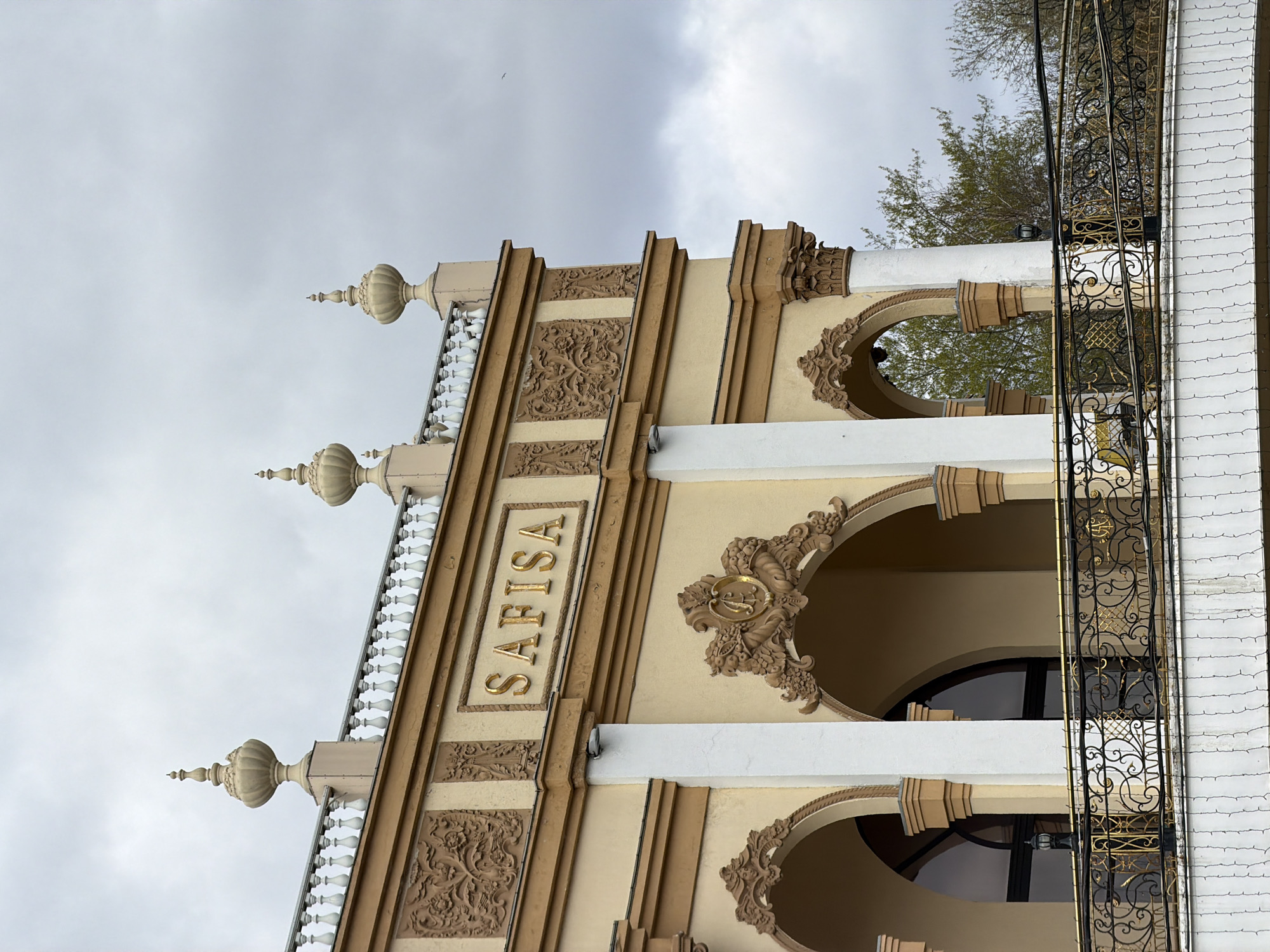 The image shows a building facade in Moscow, Russia, with the name "SAFISA" prominently displayed. The building exhibits a blend of architectural styles, possibly reflecting historical influences. The facade features decorative elements such as ornate carvings and arched windows. A wrought-iron balcony runs along the side of the building. The sky is overcast, which suggests a cloudy day.