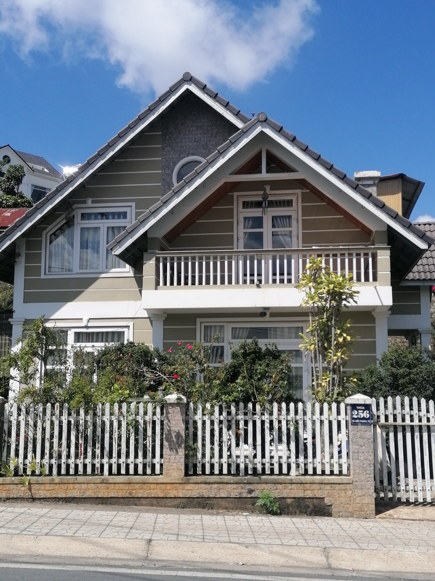The image captures a two-story house in Da Lat, Vietnam, with a distinctive gable roof clad in dark gray tiles and accented with a textured gray panel and wooden beams. The house features a light green siding with horizontal white trim, and prominent white-framed windows. A white balcony with vertical balusters adorns the second story. In front of the house, a white picket fence with stone pillars encloses a garden filled with various green shrubs and flowering plants, some with red blossoms. A street with a paved sidewalk runs along the front of the property. A blue sign with white lettering indicating the address "THUA 256" is affixed to one of the stone pillars of the fence. The sky above is a bright blue with scattered white clouds.