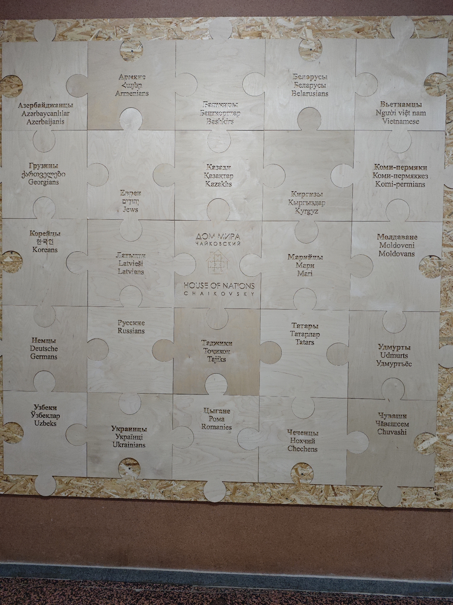 The image captures a large, light-colored wooden puzzle display mounted on a textured, rough wooden (likely OSB) backing board in an indoor setting. The puzzle comprises multiple individual pieces, each engraved with the name of an ethnic group in Russian and at least one other language, often including the group's native tongue and/or English.
At the center of the display are two puzzle pieces that read "ДОМ МИРА ЧАЙКОВСКИЙ" (House of Peace Tchaikovsky) and "HOUSE OF NATIONS CHAIKOVSKY," accompanied by a stylized house logo. Surrounding this central title, individual puzzle pieces name various nationalities and ethnic groups, such as Azerbaijanis, Armenians, Bashkirs, Belarusians, Vietnamese, Georgians, Jews, Kazakhs, Komi-Permians, Koreans, Kyrgyz, Latvians, Mari, Moldovans, Germans, Russians, Tajiks, Tatars, Udmurts, Uzbeks, Ukrainians, Roma (Gypsies), Chechens, and Chuvashi. The use of a puzzle format visually represents the idea of diverse groups forming a cohesive whole. The bottom of the image shows a dark, speckled floor or ground surface. The lighting suggests an indoor public space, possibly a community center or museum, dedicated to cultural diversity and unity.