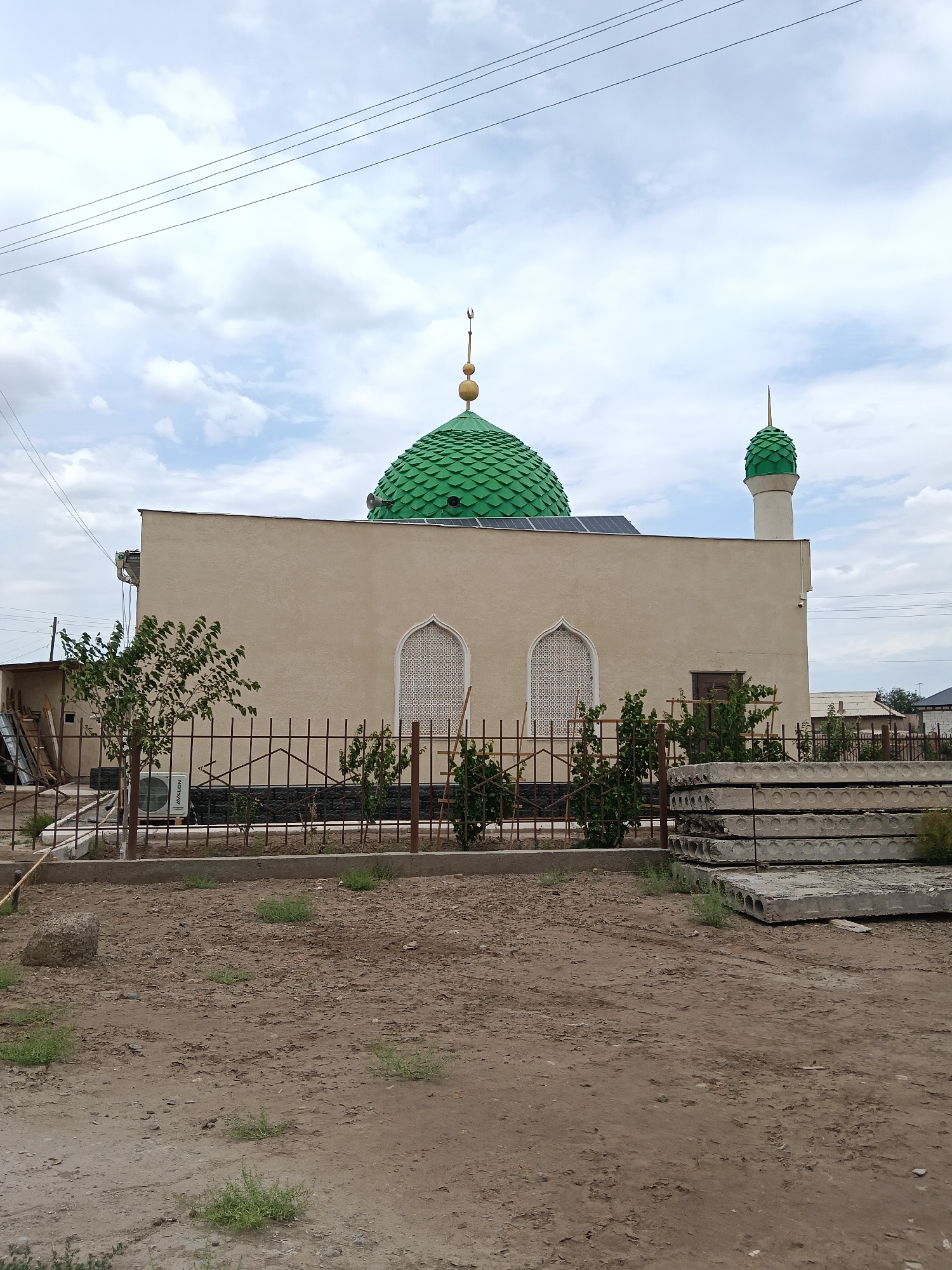 In Altinkul, Uzbekistan, a beige mosque stands prominently under a cloudy sky. The mosque features a large, green, scaled dome with a golden finial, and a smaller green dome atop a minaret. Two arched windows with intricate white latticework adorn the building's facade. In front of the mosque, a low metal fence encloses a small patch of greenery. To the left of the mosque, a small tree stands, and an outdoor air conditioning unit is visible. The foreground is a dirt lot with sparse vegetation and a stack of large, circular concrete slabs. Overhead, power lines cross the sky.