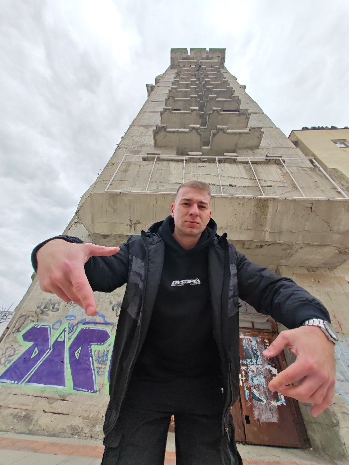 A single adult male is depicted centered in the foreground, standing before the distinctive unfinished concrete high-rise building, commonly known as the "Nedostroen blok," in Shumen, Bulgaria. The subject has short, light-colored hair and wears a black hooded sweatshirt with "DYSTOPIA" text, a black puffer jacket with dark patterned upper sections, black trousers, and a silver-toned wristwatch on his left wrist. He extends both arms forward and slightly downward, with open hands and curled fingers, looking directly at the camera with a neutral expression. The multi-story concrete structure behind him features numerous identical, staggered concrete balconies or protrusions along its vertical facade. The lower part of the building to the left displays purple and light green graffiti. To the subject's right, a rusted metal door or panel is visible, covered with torn posters or markings. The sky is overcast, and a partial view of another light-colored building is seen on the far right edge of the frame.