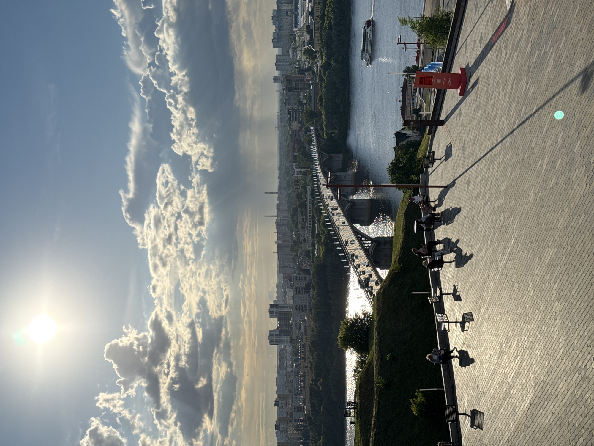 The image captures a panoramic view of a cityscape from an elevated perspective, likely during late afternoon or early evening due to the long shadows and the warm glow of the sun. The scene is dominated by a wide river, with a bustling bridge spanning across it, carrying vehicular traffic. On the far bank of the river, a dense urban landscape unfolds, characterized by numerous buildings of varying heights, including modern high-rises. Lush green trees line the riverbank, creating a contrast with the concrete structures.
In the foreground, a paved walkway with a brick-like pattern stretches across the frame, casting long shadows from a red directional sign and lampposts. Several benches are visible along the walkway, with a few people seated on them, appearing relaxed and enjoying the view. The presence of people suggests a public space, possibly a park or a promenade, designed for leisure and recreation.
The sky is a striking element, with a bright sun shining through scattered clouds that exhibit a dramatic, almost painterly quality. The sunlight glints off the river's surface, creating a shimmering effect. The overall mood is one of tranquility and urban vibrancy, with the natural beauty of the sky and water juxtaposed against the man-made environment.
While no specific text is clearly legible on the objects, the red directional sign suggests it provides information or guidance within the public space. The location context of Nizhny Novgorod, Russia, helps to place this scene along the Volga River, a significant waterway in the region. The weather appears pleasant, with clear skies and sunshine, contributing to the inviting atmosphere of the scene.
