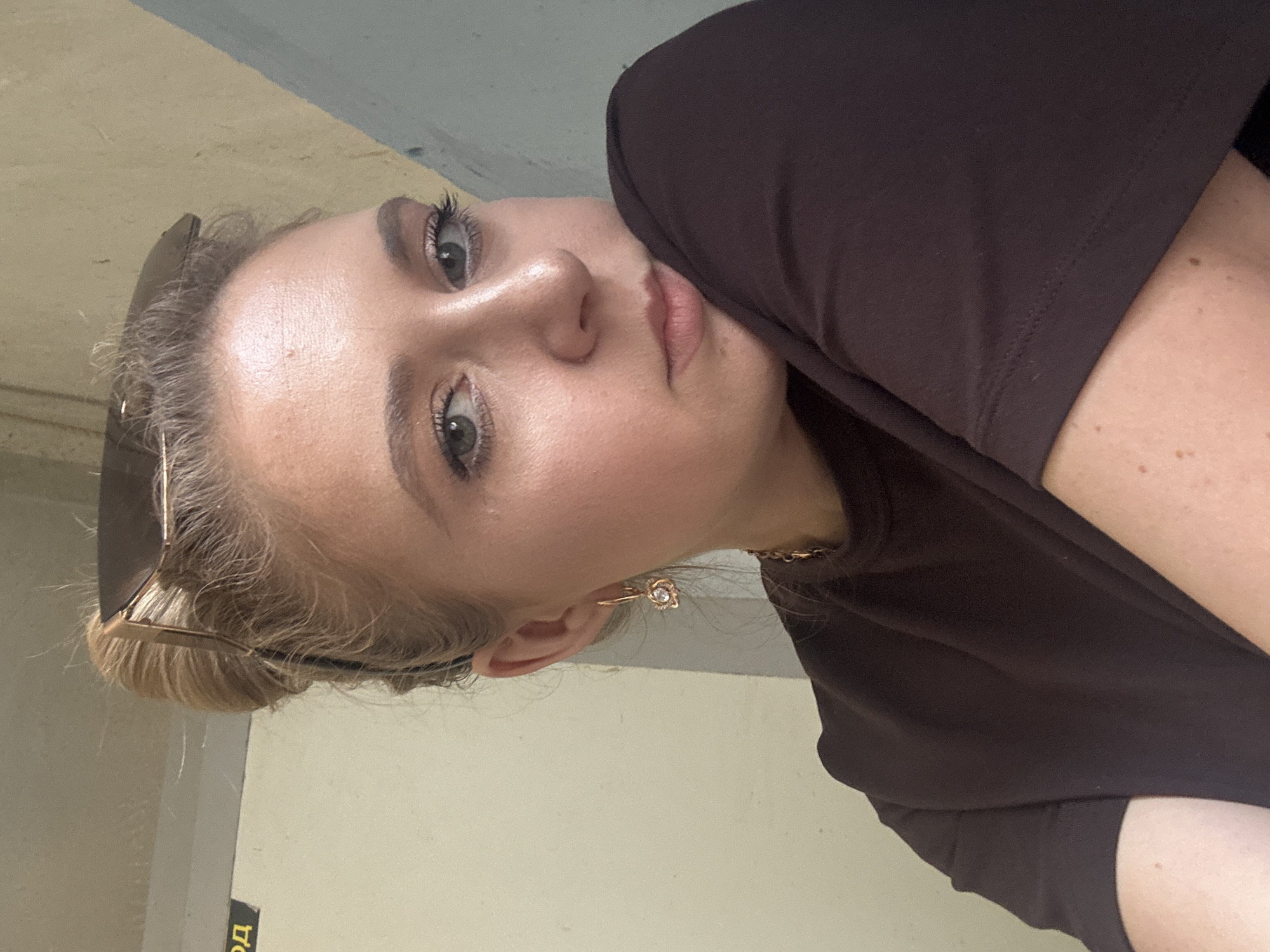 A young woman with blonde hair pulled into a bun and sunglasses perched on top of her head is captured in a close-up, slightly angled selfie. She is wearing a dark brown t-shirt and a gold chain necklace. Her makeup includes mascara, eyeshadow, and lip gloss. A diamond earring dangles from her left ear. The background consists of a light-colored wall with some writing on it and a darker, textured wall. The lighting is natural and casts a soft glow on her face. She is looking directly at the camera with a neutral expression.