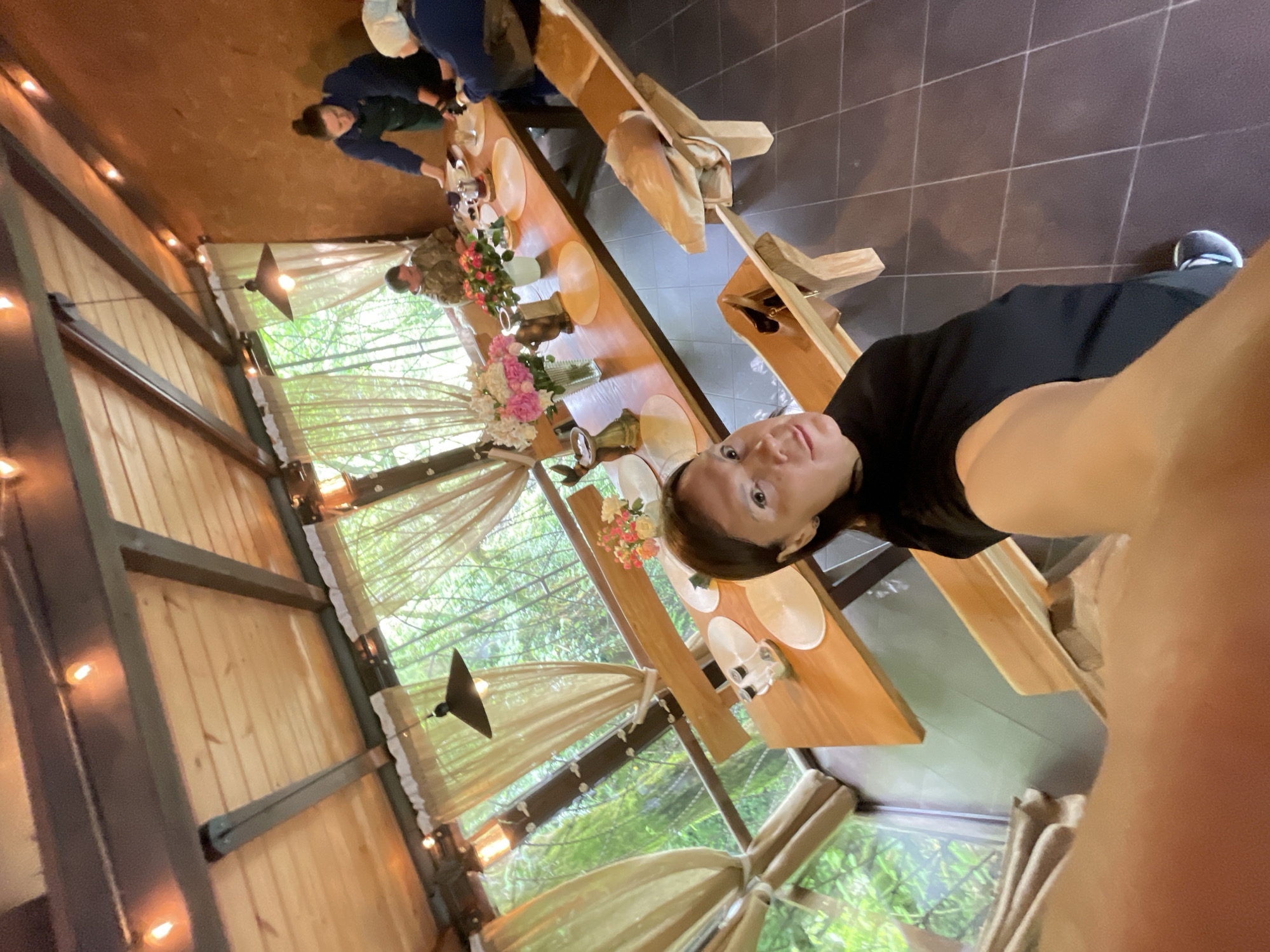 Captured in Solokh-Aul, Russia, the image shows a woman taking a selfie in what appears to be an outdoor dining area. The area is decorated with draping fabric, lights, and flower arrangements. A long wooden table is set with plates and glasses, indicating a meal or gathering is in progress. Other individuals can be seen in the background, seemingly engaged in activity related to the setting of the table.