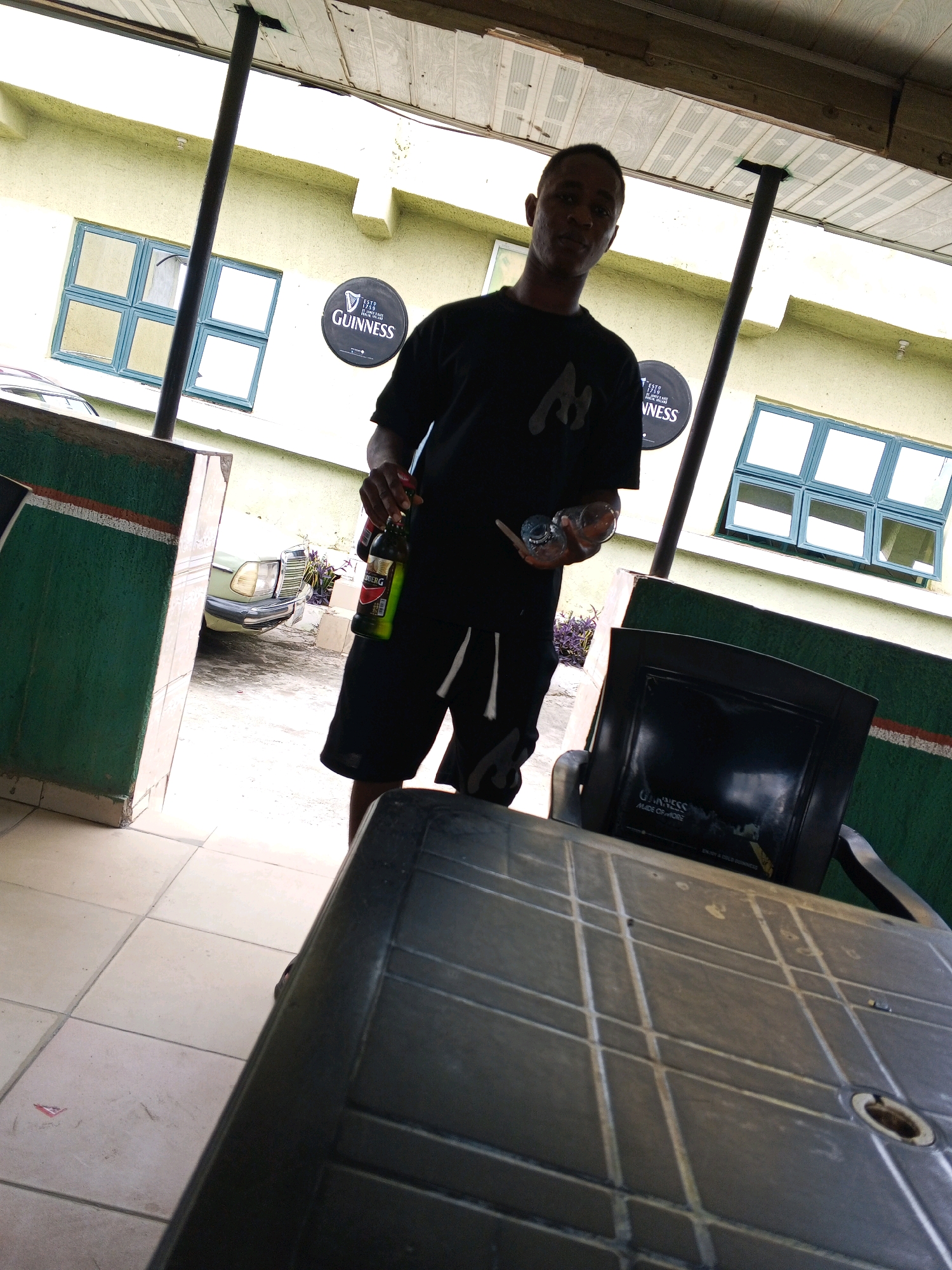 A young Black male is standing under an overhang in what appears to be an outdoor seating area of a bar or restaurant, likely in Zango-ndaji, Nigeria. The man is wearing a black t-shirt and black shorts with white stripes. He is holding a green bottle of Guinness in his right hand and two small glasses in his left. Behind him, the light-colored building has several windows with blue frames and a circular Guinness sign. A light-colored car is parked to the left of the man, and a black plastic table and chair are in the foreground. The ground is tiled.