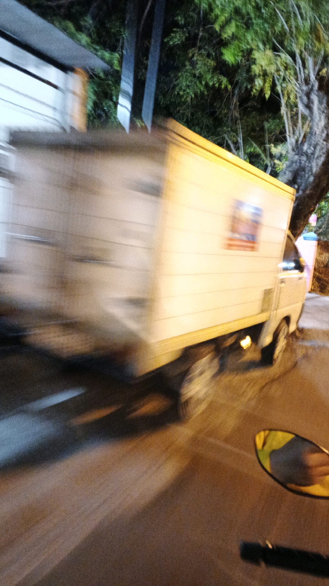The image depicts a blurred motion shot of a white delivery truck with a cargo box, moving from left to right. The truck has a yellow stripe along its bottom edge and a logo or sticker on its side. The scene appears to be at night, with artificial lighting illuminating the truck and casting shadows on the asphalt road. Trees and foliage are visible in the background, suggesting an outdoor urban environment. The photograph was taken from a moving vehicle, possibly a motorcycle, as indicated by the presence of a side mirror in the lower right corner reflecting a yellow object. The location is identified as Surakarta, Indonesia.