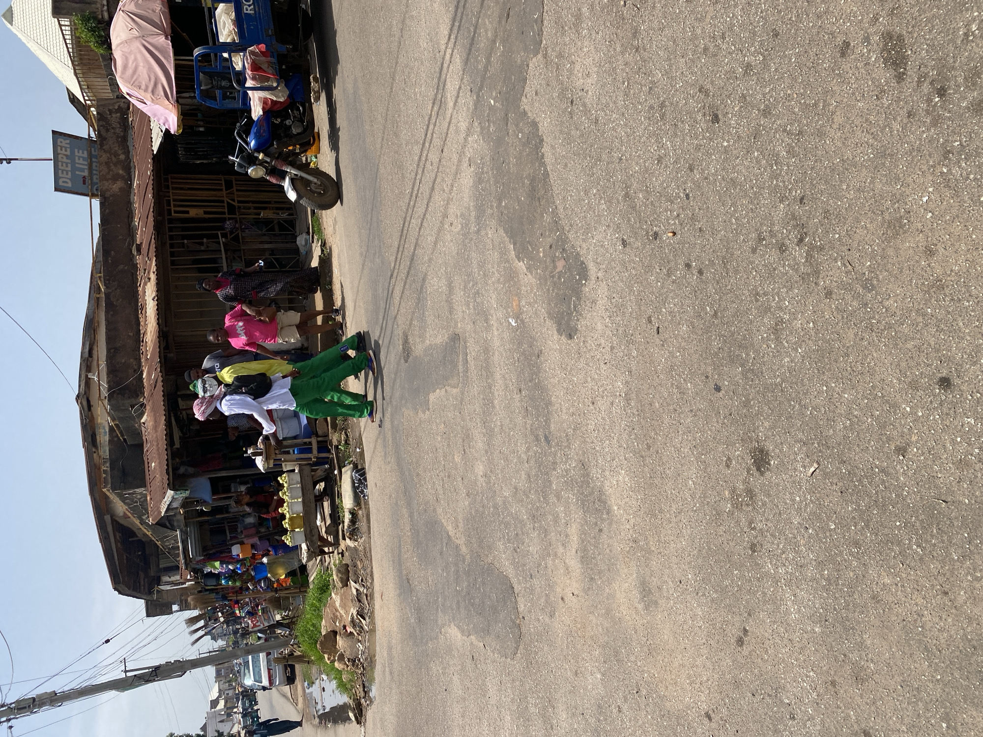 This is a street scene in Jos, Nigeria, on a bright, sunny day. The foreground is an asphalt road with visible patches and some water puddles. On the right side of the road, a blue three-wheeled vehicle, possibly a tricycle, is parked. Beyond the road, a row of market stalls and shops line the street. The buildings appear weathered with corrugated metal roofs. A sign that reads "DEEPER LIFE" is visible above one of the structures. Several people are present in the scene, some walking and others standing near the market stalls. One group of individuals in the mid-ground are wearing green pants and white shirts, with one person also wearing a white head covering. Another woman in a pink shirt and shorts is visible near the tricycle. The market area is stocked with various goods, including what appear to be brooms and buckets. The overall impression is a bustling, open-air market in an urban environment.