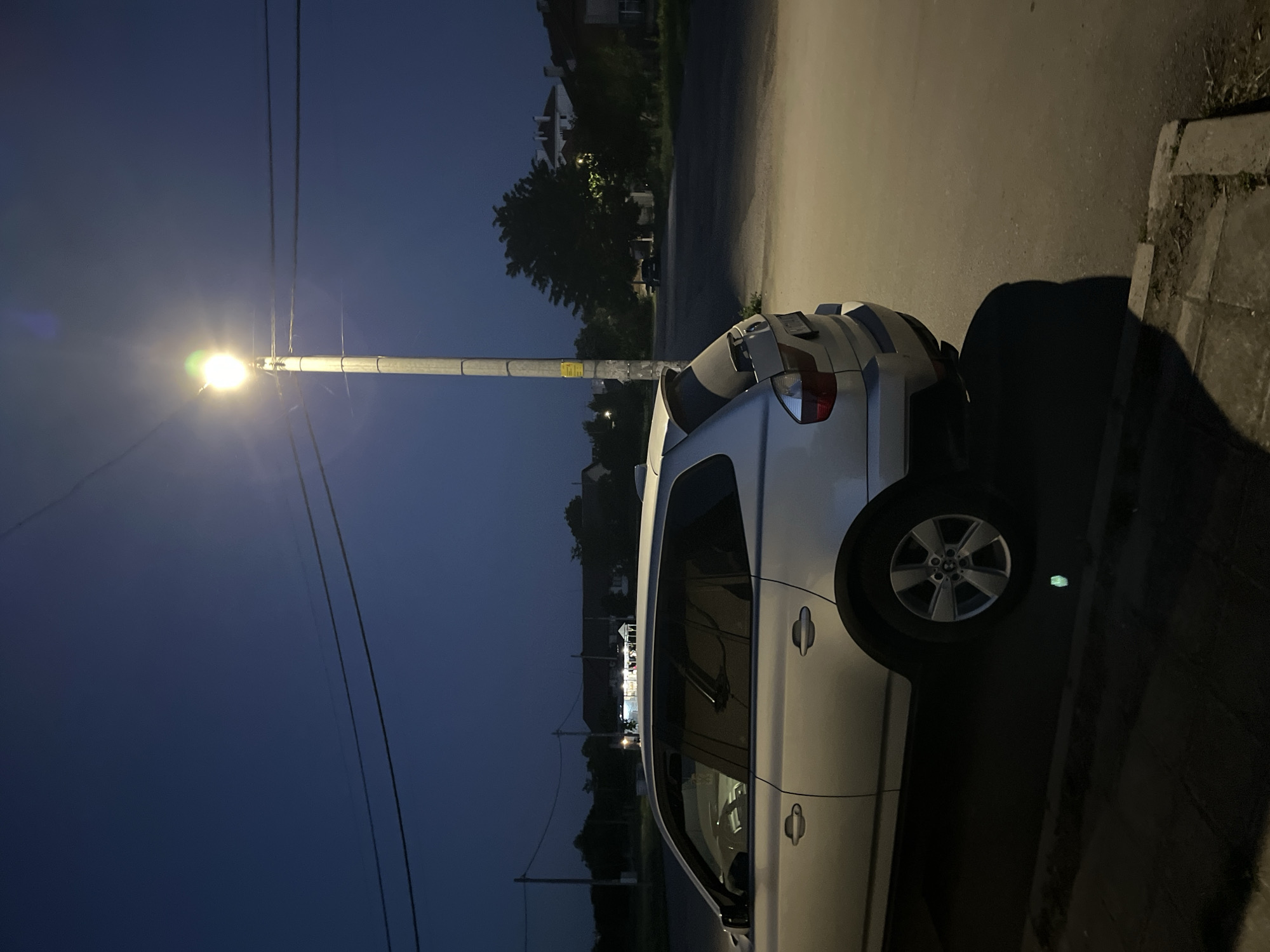 A silver car is parked on the side of a street in what appears to be the city of Elhovo, Bulgaria, based on the street signs. The sky is dark, suggesting it is nighttime. A street lamp is lit, illuminating the car and the immediate area. The car is positioned close to the curb. The surrounding environment includes trees, buildings, and a sidewalk. The scene is still and quiet, with no visible human activity.