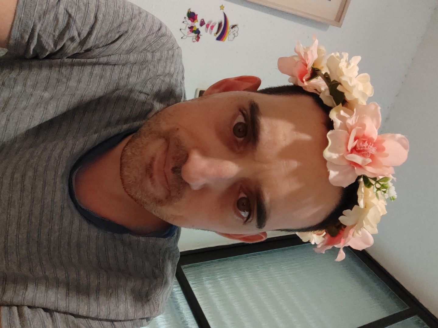 A man with dark hair and light skin, wearing a grey shirt and a flower crown of light pink and white flowers, poses for a photo. The background is a white wall with a colorful sticker on it and a window with frosted glass. The image is rotated. There is no location or city specified in the image.