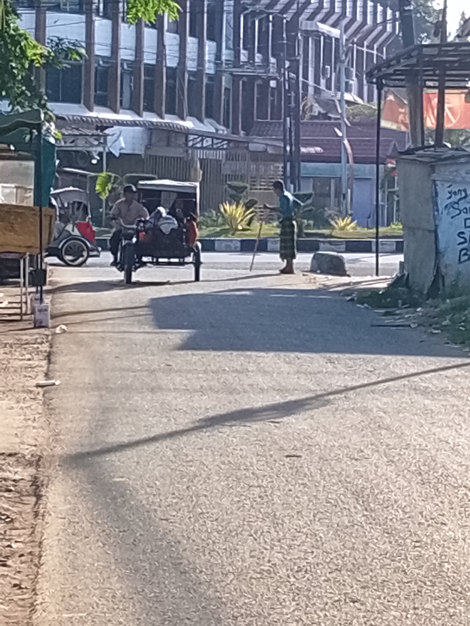 An outdoor, eye-level, medium shot captures a street scene in Lhokseumawe, Indonesia. In the foreground, a grey asphalt road occupies most of the frame, with a faint shadow of a tree stretching across it. To the left, a portion of a building with a wooden facade is visible, along with a grey metal table.
In the mid-ground, a motorized tricycle, known locally as a becak, is the central focus. A man wearing a light brown shirt and a dark cap rides the becak, which has a sidecar carrying two passengers, one in red and one in black. To the right of the becak, another man stands on the sidewalk, dressed in a blue shirt and a patterned sarong, holding a long stick. He appears to be facing away from the camera, possibly observing the becak.
The background reveals a row of multi-story buildings with a modern architectural style, featuring prominent vertical elements and a patterned facade. A street light pole is also visible, along with some vegetation and a concrete wall with faded blue and white writing on it. The lighting suggests it is daytime, with the sun casting distinct shadows.
