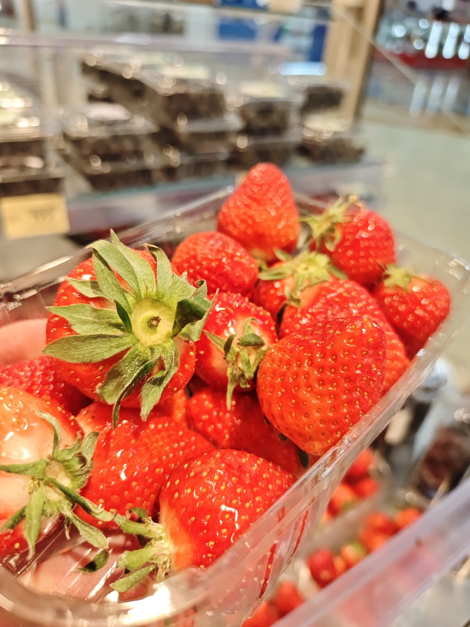 A close-up shot focuses on a clear plastic container filled with fresh strawberries. The strawberries are bright red, with green stems attached. In the background, blurred rows of other packaged food items are visible in a display case. The strawberries are in what appears to be a store in Yekaterinburg, Russia.