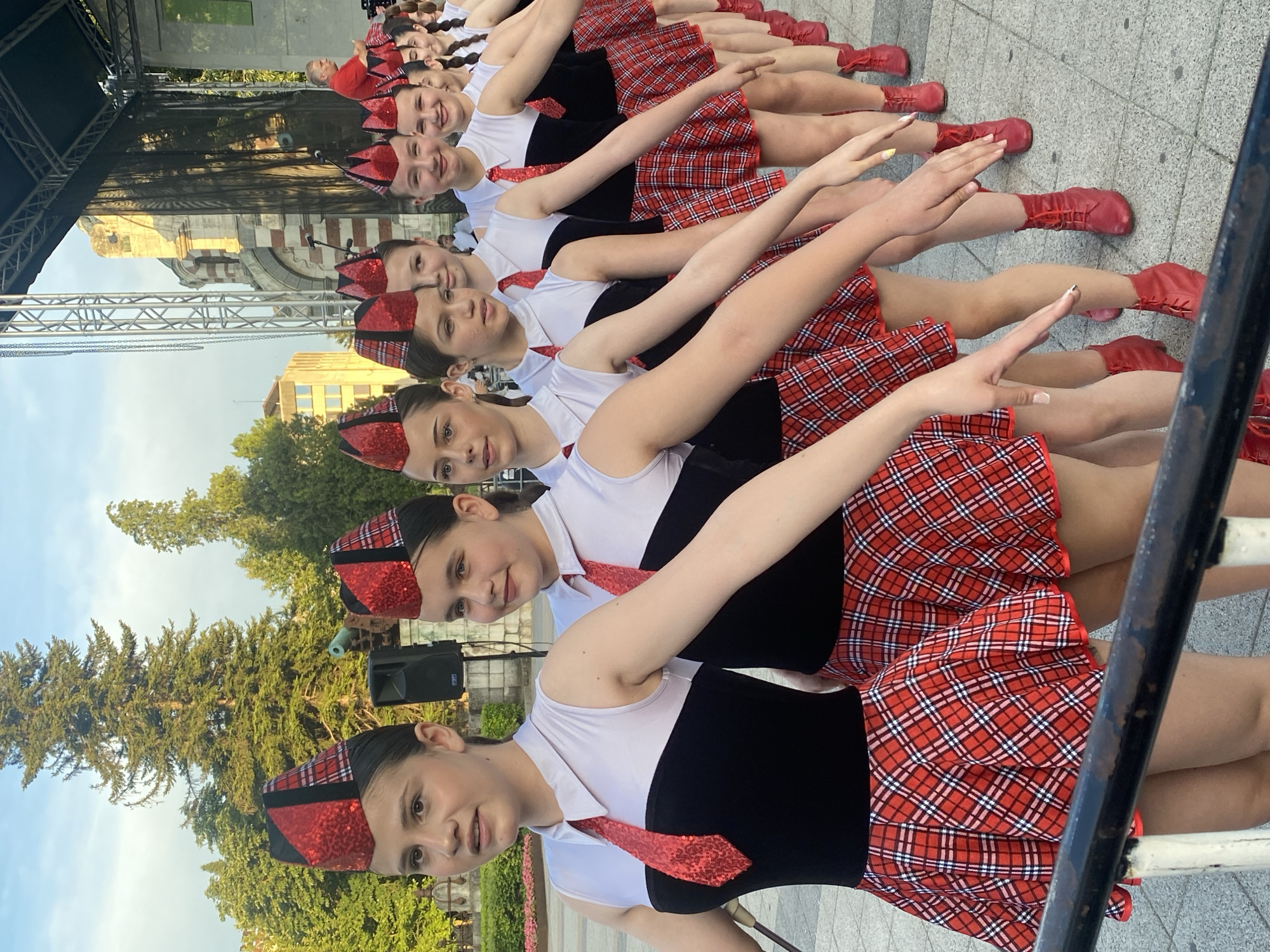A line of young women, likely performers, are positioned outdoors in a city, specifically Pleven, Bulgaria, as indicated by the context. They are dressed in matching uniforms consisting of red and black plaid skirts, white tops with black sleeveless vests, and red sequined hats adorned with plaid accents. Each performer also wears a red sequined tie. The women are arranged in a diagonal line, facing forward and to the left of the frame. Their hands are extended forward in a coordinated gesture, suggesting they are in the middle of a performance or a posed photograph. The ground is paved with grey cobblestones. In the background, trees, a stage structure with lighting equipment, and architectural buildings are visible, illuminated by natural daylight, likely late afternoon or early evening given the soft lighting. A railing runs along the bottom edge of the image.