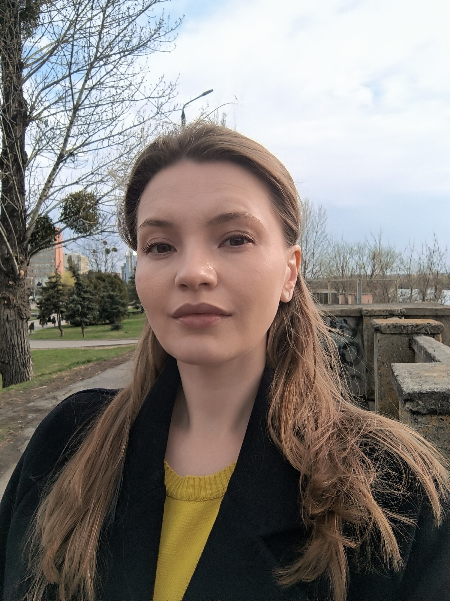 A medium-shot photograph shows a woman with light skin and long, light brown hair posing for a selfie. She wears a yellow sweater beneath a dark coat. Behind her, to the left, is a bare tree with several mistletoe clumps in its branches and a park with green grass and evergreen trees. A building is visible in the background. To the right, there is a concrete railing. The sky is overcast. Given the background elements, this photo was likely taken in Vinnytsia, Ukraine, at a park or embankment area.