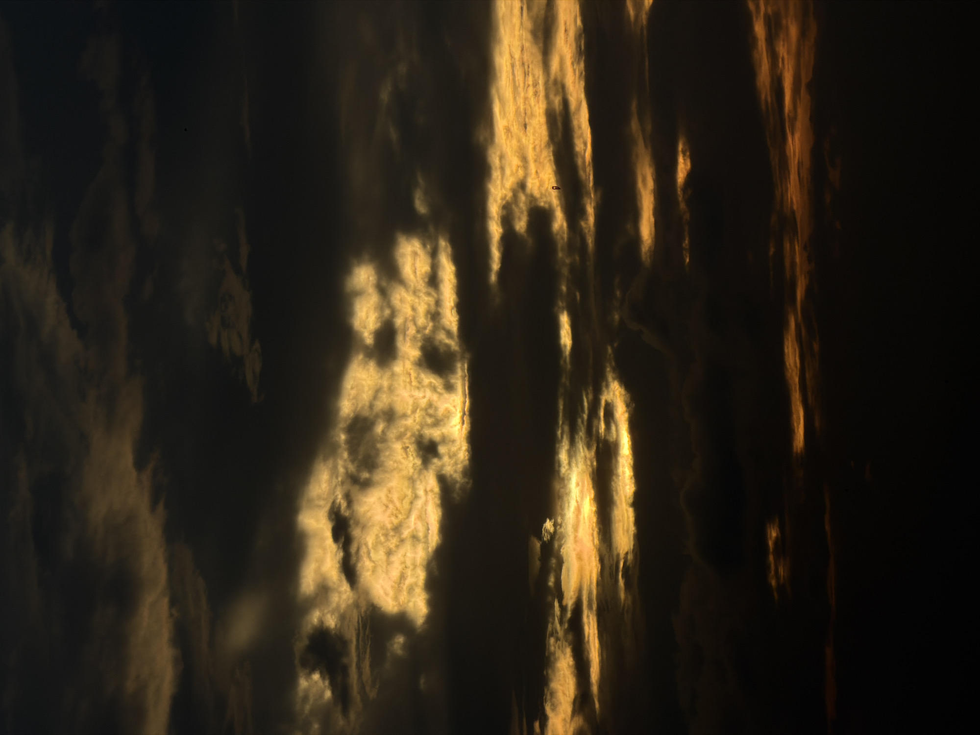 The image shows a dark and cloudy sky with streaks of golden light breaking through. The scene is characterized by dramatic lighting and textures, with heavy, shadowed clouds contrasting with brighter, illuminated areas. The lower portion of the image fades into deep darkness, suggesting the foreground or horizon is obscured. The overall impression is one of a brooding or intense sky, possibly during a sunrise or sunset. Without further information or visual cues, the specific location within North Kuta, Indonesia, cannot be determined from the image content alone.