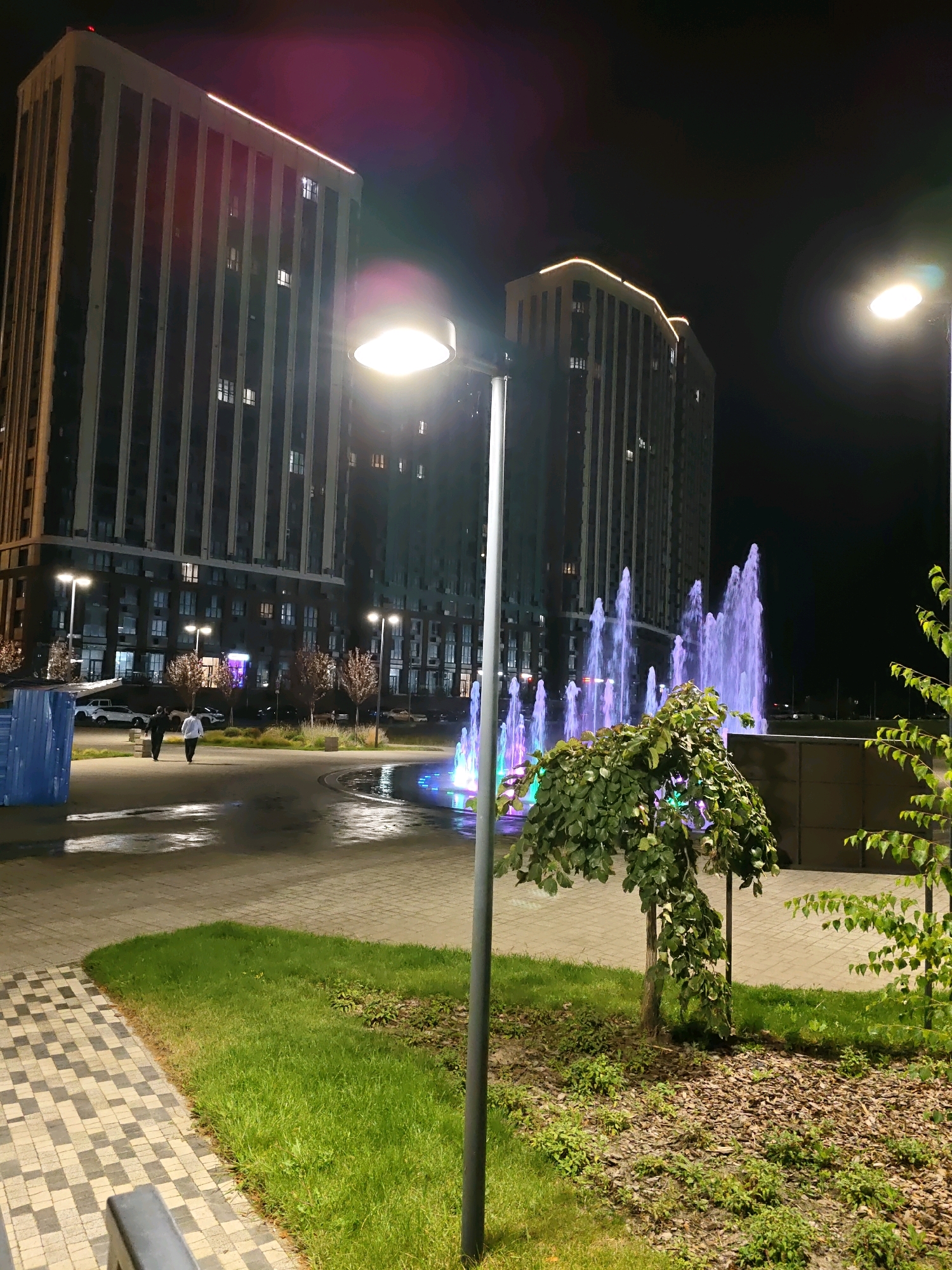 The image depicts a modern urban scene at night, likely in Rostov-on-Don, Russia, featuring two large, multi-story apartment buildings with illuminated windows. In the foreground, a well-lit pathway made of checkered pavers leads towards a fountain with vibrant purple and green lights. Two individuals, one in a dark jacket and the other in a white hoodie, are walking away from the camera on the wet pavement near the fountain. Several streetlights illuminate the area, and the ground surrounding the fountain is covered in mulch and grass, with a small, leafy tree in the center. The overall atmosphere is one of urban nighttime ambiance, with reflections on the wet surfaces adding to the visual appeal.