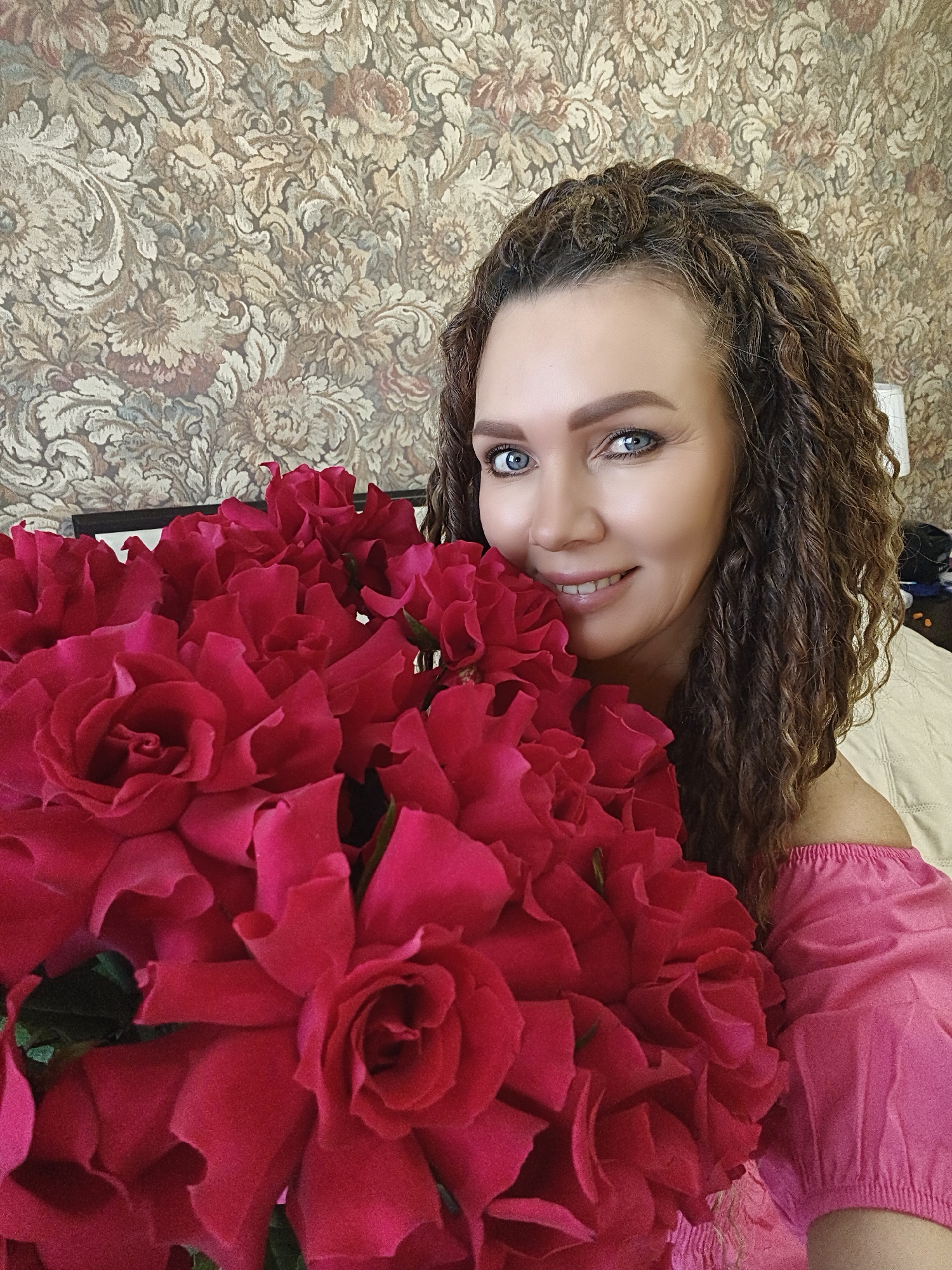 A woman is holding a large bouquet of red roses. She has curly brown hair, blue eyes, and is wearing a pink off-the-shoulder top. The background features a patterned wallpaper with floral and abstract designs. The location appears to be indoors, possibly a home or room in the city of Unknown, Unknown. The focus is on the woman and the roses she is holding.