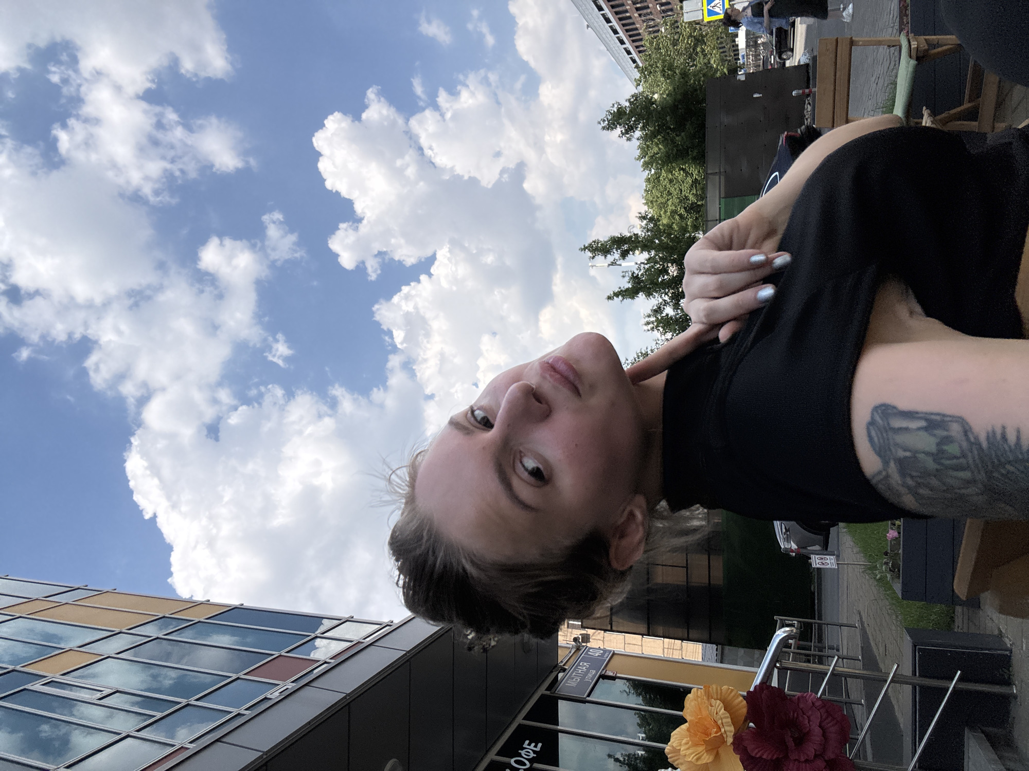 A young woman with blonde hair pulled back in a bun takes a selfie outdoors in front of a modern building with large windows. She is wearing a black top and has a tattoo on her arm. The sky is blue with fluffy white clouds. In the background, there are trees, a street, and other buildings. A sign with Russian text and the number "40" is visible on the building. Flowers are placed on a railing to the left of the woman.