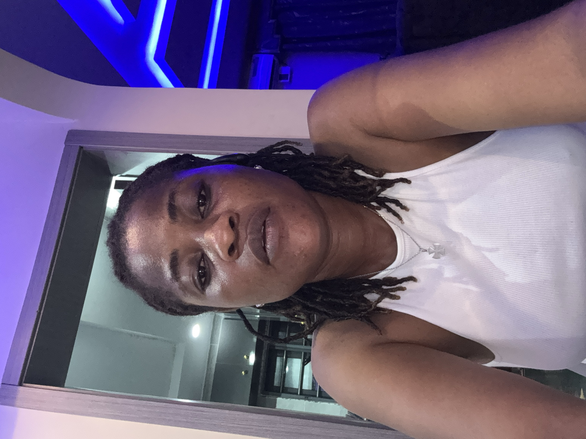 A medium shot shows a woman from the chest up, facing the camera. She has dark skin and her hair is styled in dreadlocks. She is wearing a white tank top with a silver cross necklace. Her expression is neutral. The background appears to be indoors, with purple lighting on the ceiling and blue lighting on the right side. There is a doorway or partition to the left with a wooden frame. The scene is likely a home or a commercial establishment. No specific location in Ajara, Nigeria is identifiable from the image.