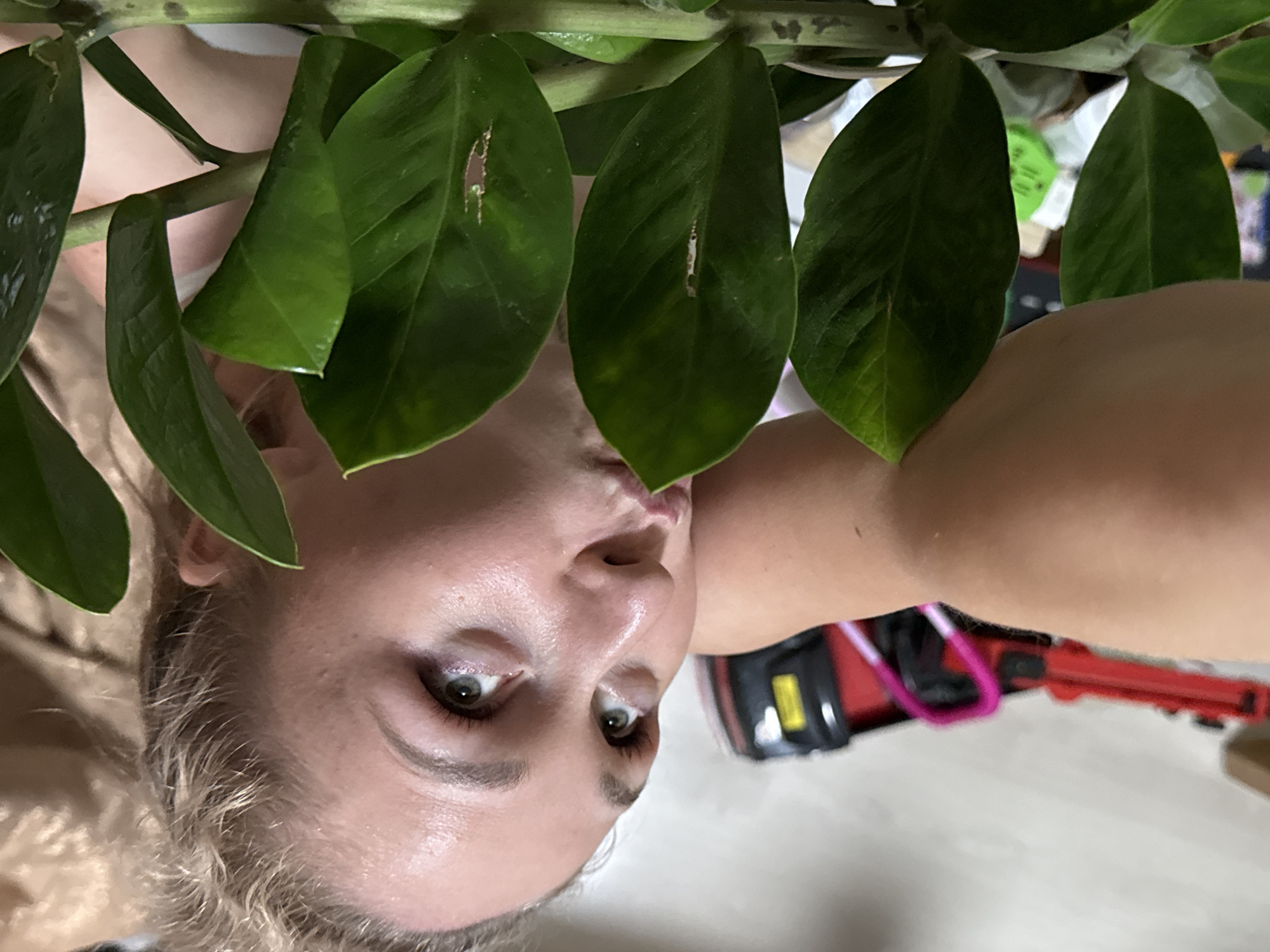 A woman with blonde hair and fair skin is positioned behind a plant with green, oval-shaped leaves. She is looking directly at the camera with a slight pout. A red and black object, possibly a toy or tool, is partially visible in the lower right corner. The setting appears to be indoors, with a light-colored floor and additional objects blurred in the background. The image does not provide any location information related to Chaikovsky, Russia.