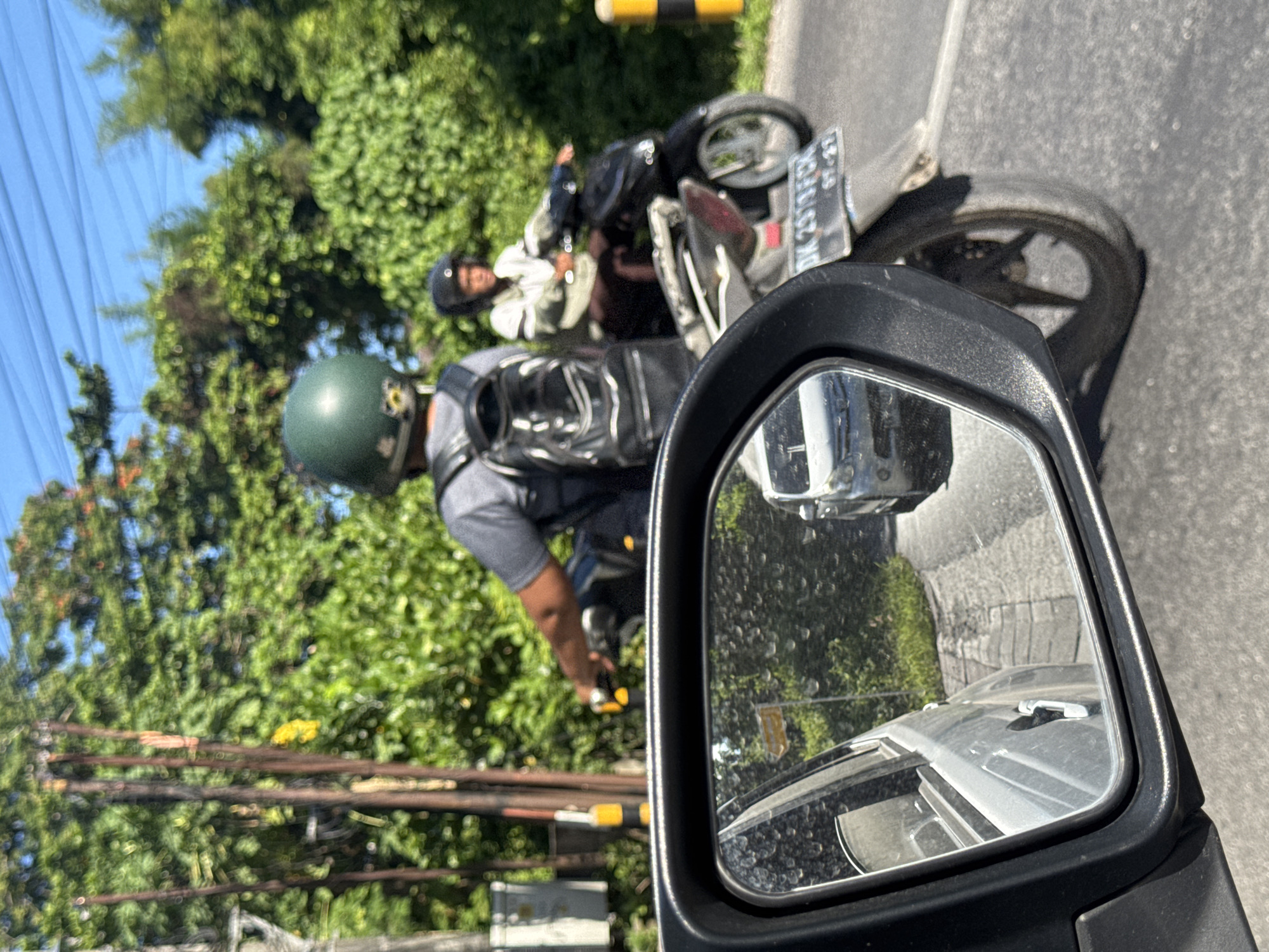 The image is a close-up shot taken from the perspective of a driver, showing the side mirror of a vehicle. Reflected in the side mirror are elements of the surrounding environment, including other vehicles on the road.
Outside the mirror, the primary subject is two people riding a motorcycle. The driver of the motorcycle is wearing a green helmet and a dark vest. A second person is on the back of the motorcycle wearing a black helmet. The license plate of the motorcycle is visible but illegible, with the letters "DK" in the top corner and "2595 FCK" below it. Lush green trees and vegetation are visible in the background, suggesting a tropical or subtropical location. Power lines are visible running in front of the trees.
Given the prompt specifying the location as Kuta Selatan, Indonesia, the scene likely captures a typical traffic situation in the area. The presence of motorcycles is common in Indonesian cities.