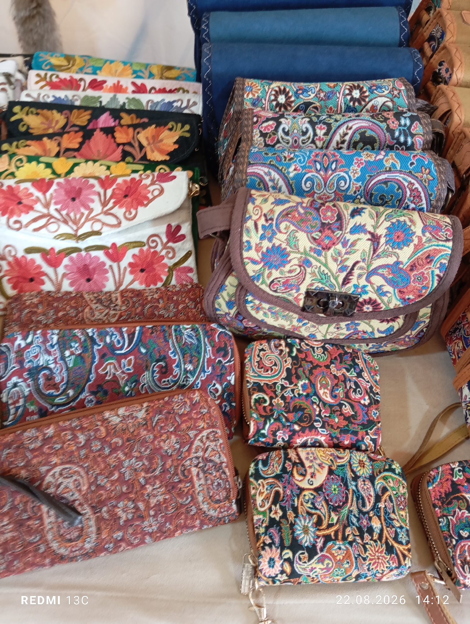 The image displays a collection of embroidered bags and clutches, likely for sale, laid out on a neutral surface. The items feature intricate patterns, including floral designs and paisley motifs in vibrant colors such as red, blue, yellow, and green. The bags are stacked and arranged in a visually appealing manner. In the background, a stack of dark blue fabric rolls is visible. The bottom of the image shows a timestamp and device model: "22.08.2026 14:12 REDMI 13C". There are no people present in the scene. The overall impression is of a market stall or a retail display of handcrafted accessories. The location in Amirsay, Uzbekistan is not discernible from the visual content of the image itself.