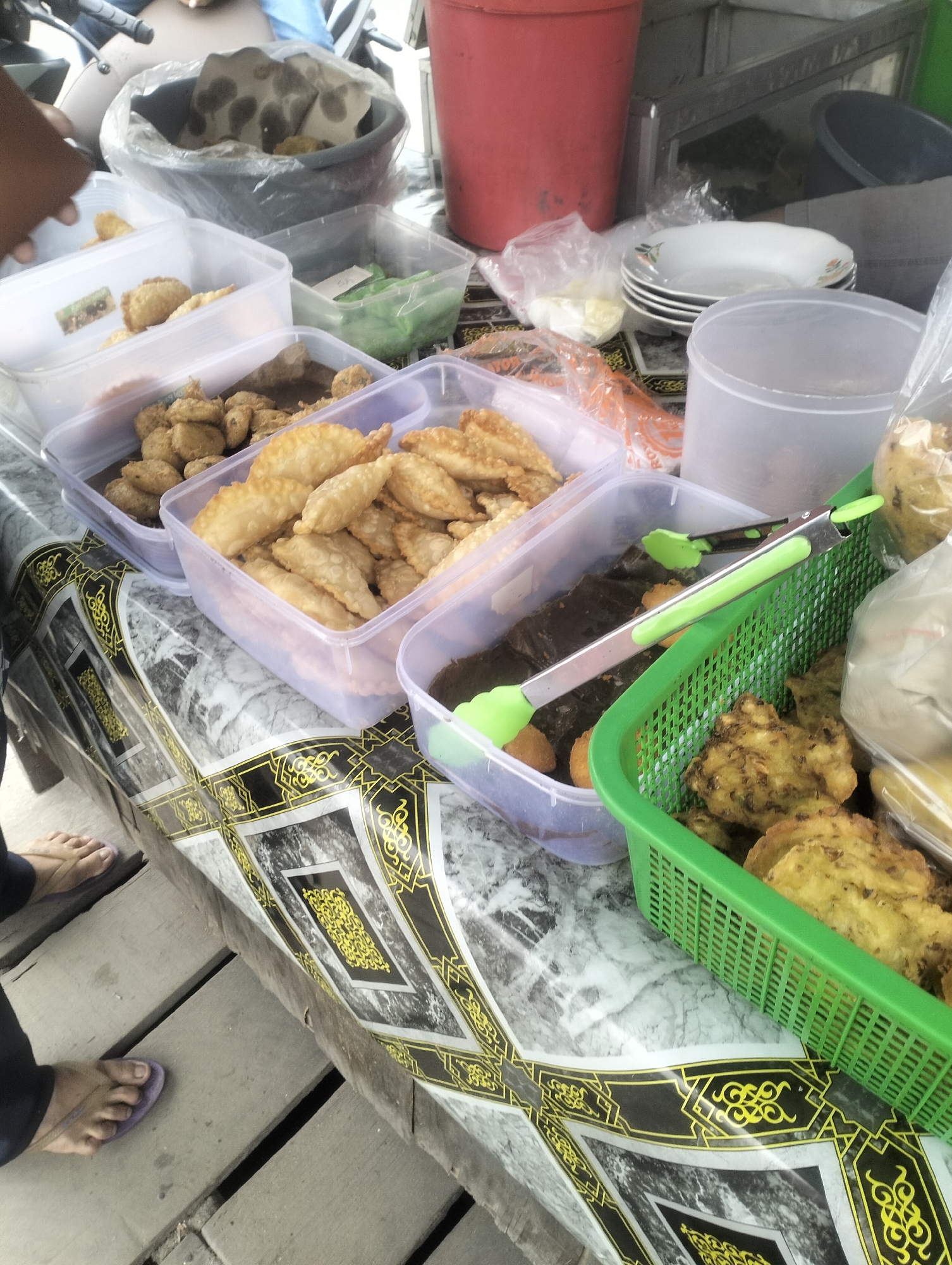 A street food stall in Dumai City, Indonesia, features an array of fried foods in clear plastic containers and a green plastic basket. In the foreground, a person's feet are visible, wearing flip-flops. The stall is set up on wooden planks, and a patterned tablecloth with black and yellow designs covers part of the surface. Behind the food, a red bucket and other containers are present. The lighting suggests it is daytime.