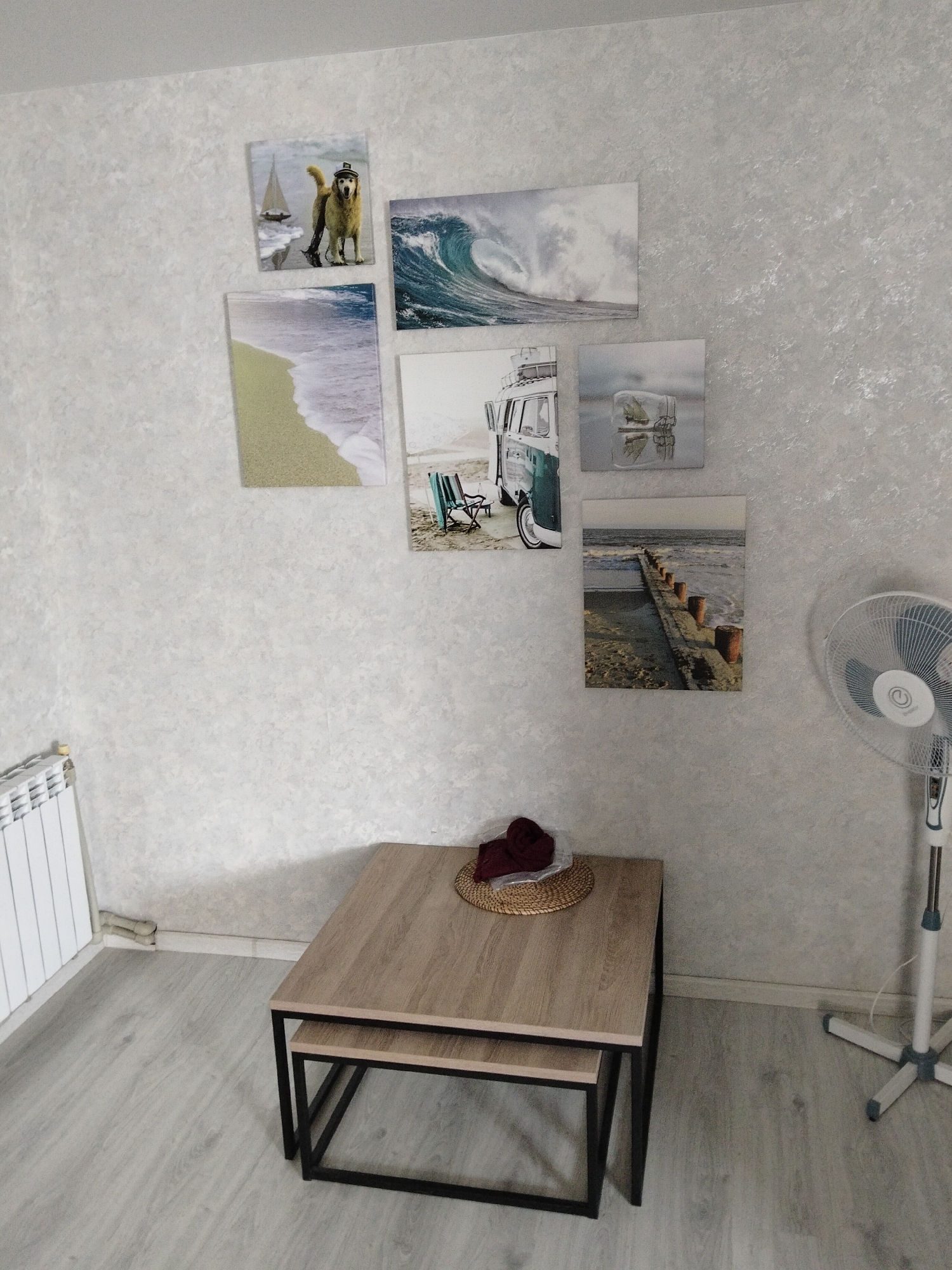 In a sparsely furnished room, likely a living area or bedroom, a stack of two nested tables with light wood tops and black metal frames sits on a light gray wood-look floor. The larger table is positioned in front of the smaller one. On the larger table, a dark red knitted item rests on a round woven mat, which is itself placed on a clear plastic bag.
The wall behind the table is textured with a light gray, almost concrete-like pattern. Several framed pictures are hung on the wall. From top left, clockwise, they depict: a golden retriever wearing a hat and standing on a beach with a sailboat in the background; a powerful ocean wave crashing; a translucent cube with a small sailboat inside, reflected in water; a wooden pier extending into the sea with cylindrical wooden posts lining its edge; a vintage green and white camper van parked on a sandy beach with two empty beach chairs in the foreground; and a sandy beach with gentle waves rolling in.
To the right of the pictures, a white oscillating pedestal fan with a black grill stands on the floor. To the left of the tables, a white radiator is mounted on the wall. The overall impression is one of a room decorated with a coastal and travel theme, with a minimalist aesthetic. The image was taken in Novinki, Russia.