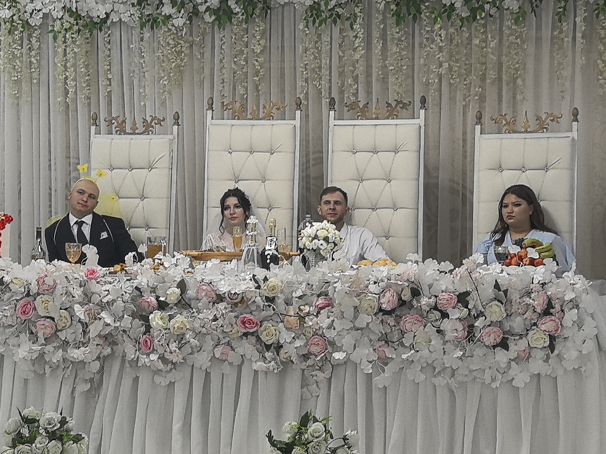 The image shows a wedding reception with four people seated at a decorated table. The table is covered with white fabric and adorned with an elaborate floral arrangement of white, pink, and cream roses. Behind the table, a white curtain serves as a backdrop, with hanging white floral garlands and green leaves.
From left to right, the individuals are:
1. A man with a shaved head, wearing a black suit, white shirt, and black tie. He has a yellow accessory on his head.
2. A woman with dark hair, wearing a white dress. She is looking slightly to her left.
3. A man with short brown hair, wearing a light-colored shirt. He is looking forward.
4. A woman with long dark hair, wearing a light blue top. She is looking to her right.
On the table, there are glasses, bottles, and a fruit arrangement. The people appear to be seated and posing for a photograph. The overall scene suggests a formal event, likely a wedding ceremony or reception, taking place indoors. The lighting is soft and diffused, illuminating the subjects and decor.