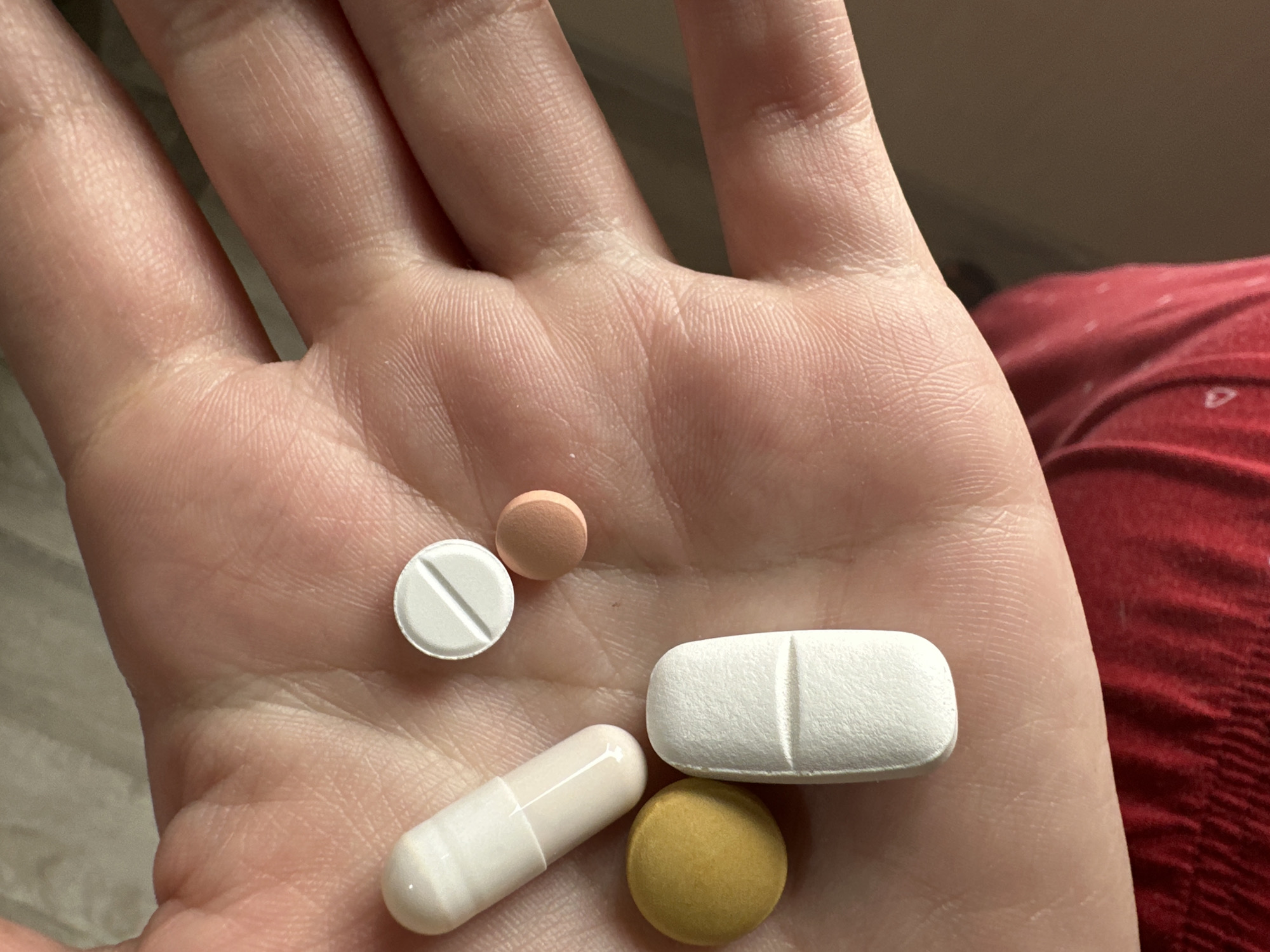 A human hand is holding five pills of different shapes, sizes, and colors. There is a white capsule, a white round tablet with a score, a peach-colored round tablet, a large white oblong tablet with a score, and a yellow round tablet. The hand is light-skinned. The background includes blurred elements of what appears to be clothing. There is no indication of a location in the city of Perm, Russia.