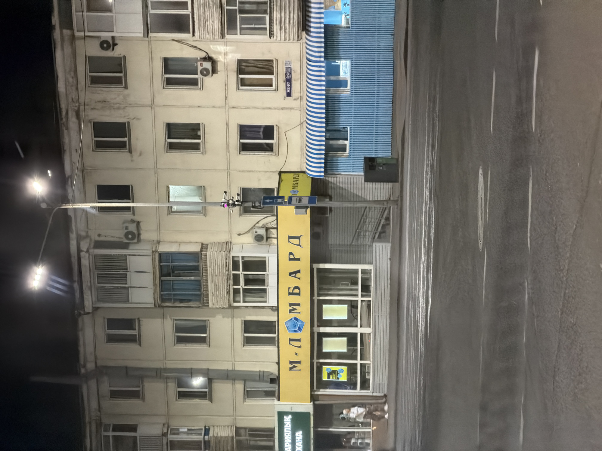 The image shows a street scene at night in Astana, Kazakhstan. The central focus is a building with a bright yellow awning that reads "М-ЛOMБAPД" with a blue diamond logo. Below the awning is a storefront with large glass windows, displaying two illuminated square panels. To the left of the yellow awning is a green sign that is partially visible, reading "АРЫЛЫҚ...ХАНА". To the right of the yellow awning is a blue structure with white striped awnings, featuring several windows. A street lamp with bright lights is visible in the upper portion of the image, illuminating the building and street. There are security cameras mounted on a pole near the storefront. A trash bin is located next to the blue structure. The street in the foreground appears wet and reflects the lights, suggesting it may have rained. Two individuals, a man and a woman, are visible on the sidewalk near the storefront. The building itself appears to be an apartment building with multiple stories and windows.