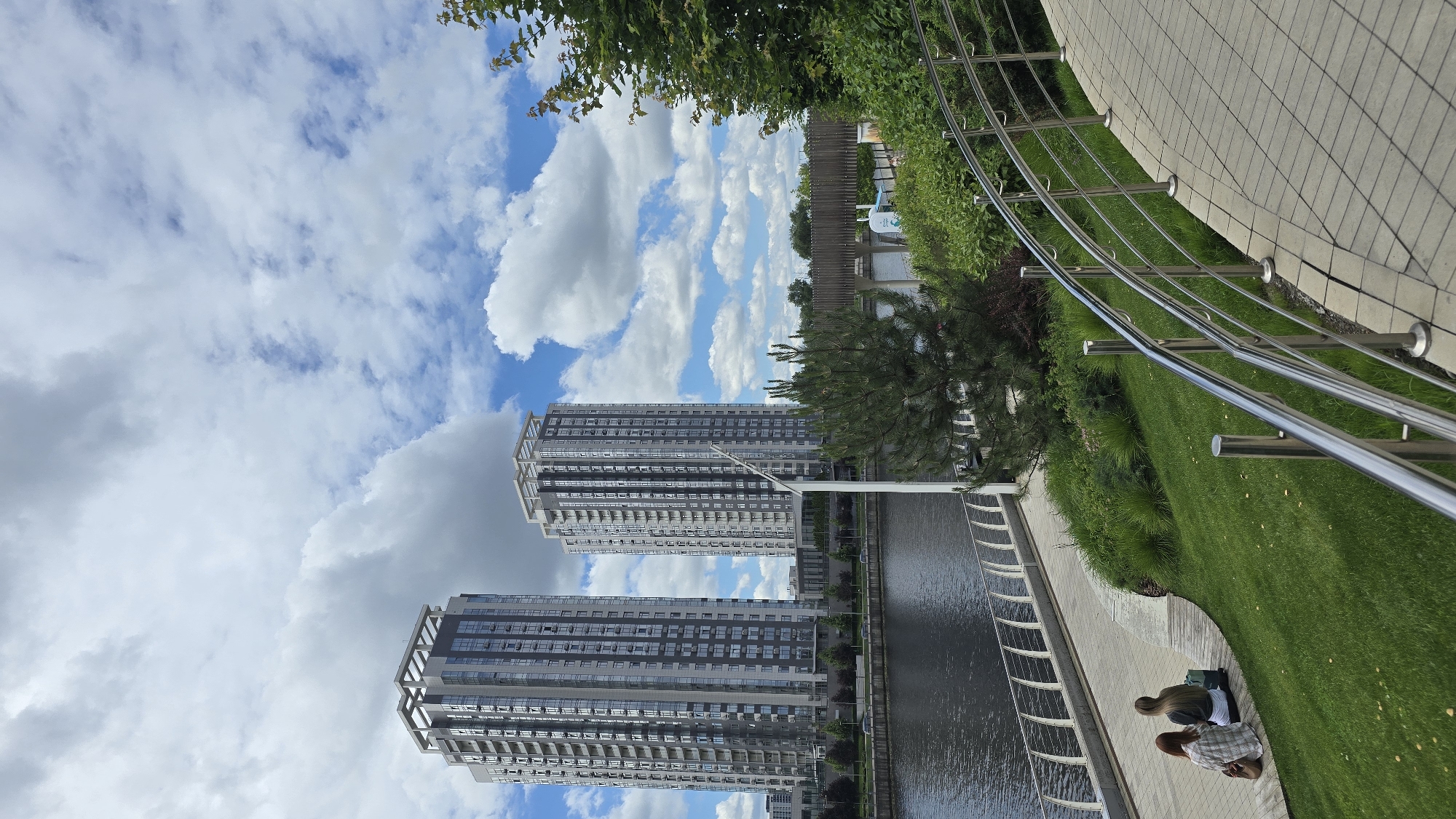 The image captures a waterside promenade in Kyiv, Ukraine, likely on a bright, cloudy day. In the background, two tall, modern apartment buildings with numerous windows dominate the skyline. A body of water, possibly a river or canal, runs along the left side of the frame, with a promenade or walkway bordering it. Two individuals, appearing to be women, are seated on a wooden deck area on the grassy embankment, facing away from the camera. One is wearing a plaid shirt and the other a dark top. A blue bag and a brown bag are also visible near them. Lush green grass and various types of vegetation, including trees and bushes, fill the middle ground and right side of the image. A metal railing lines the curved walkway in the foreground, which is paved with interlocking bricks. A pedestrian bridge can be seen in the distance, spanning the water. The sky is filled with a mix of white and gray clouds, interspersed with patches of blue.