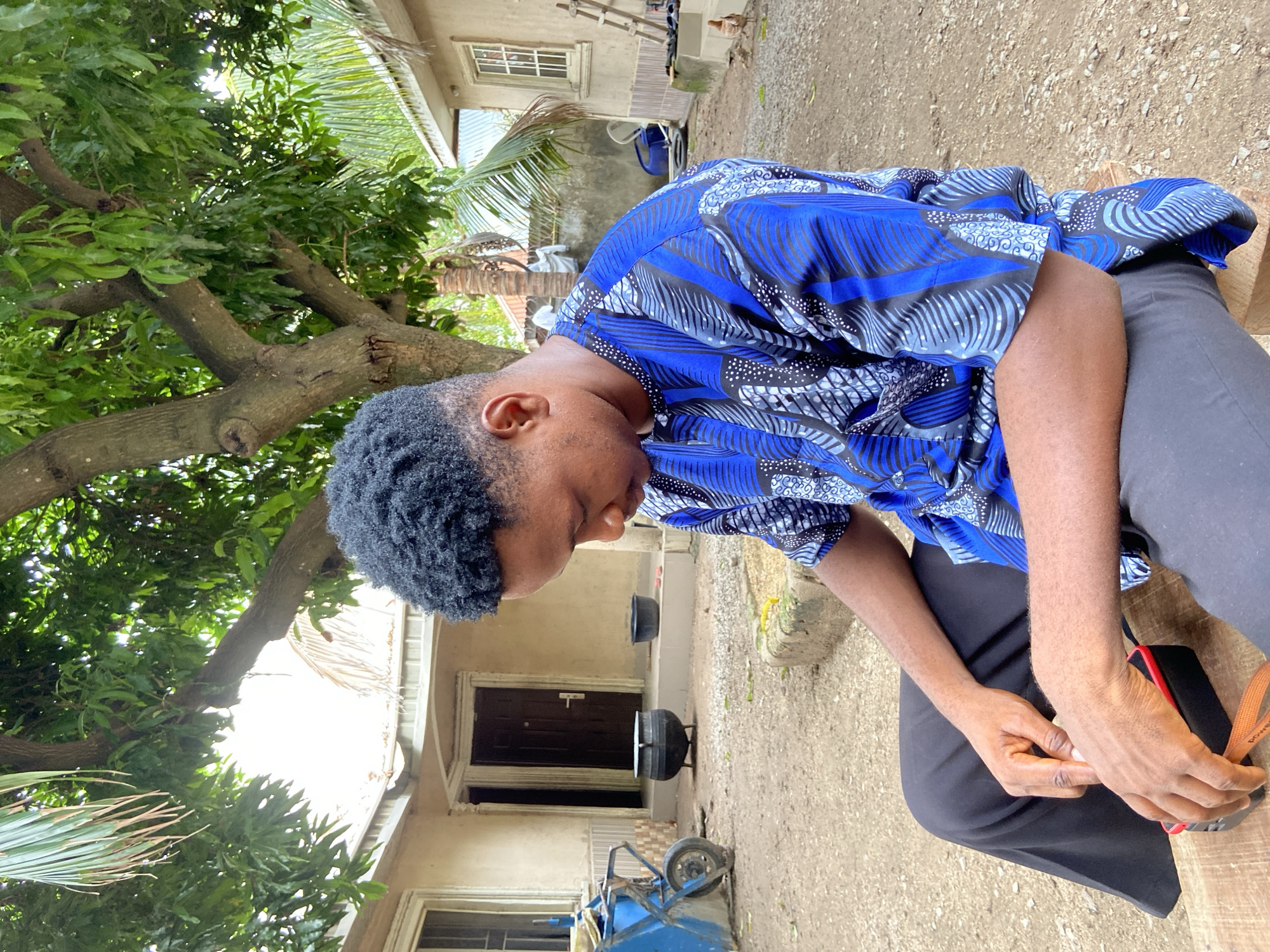 A young Black man sits outdoors, likely in Mararaba, Nigeria. He wears a blue patterned shirt and dark pants, looking down at his hands. He sits on a wooden surface, and a red and black object is visible beside him. The background includes a tree with green foliage and a building with a dark-colored door. Two black pots and a blue wheeled object are near the building. The ground is a light-colored surface, appearing to be dirt or gravel.