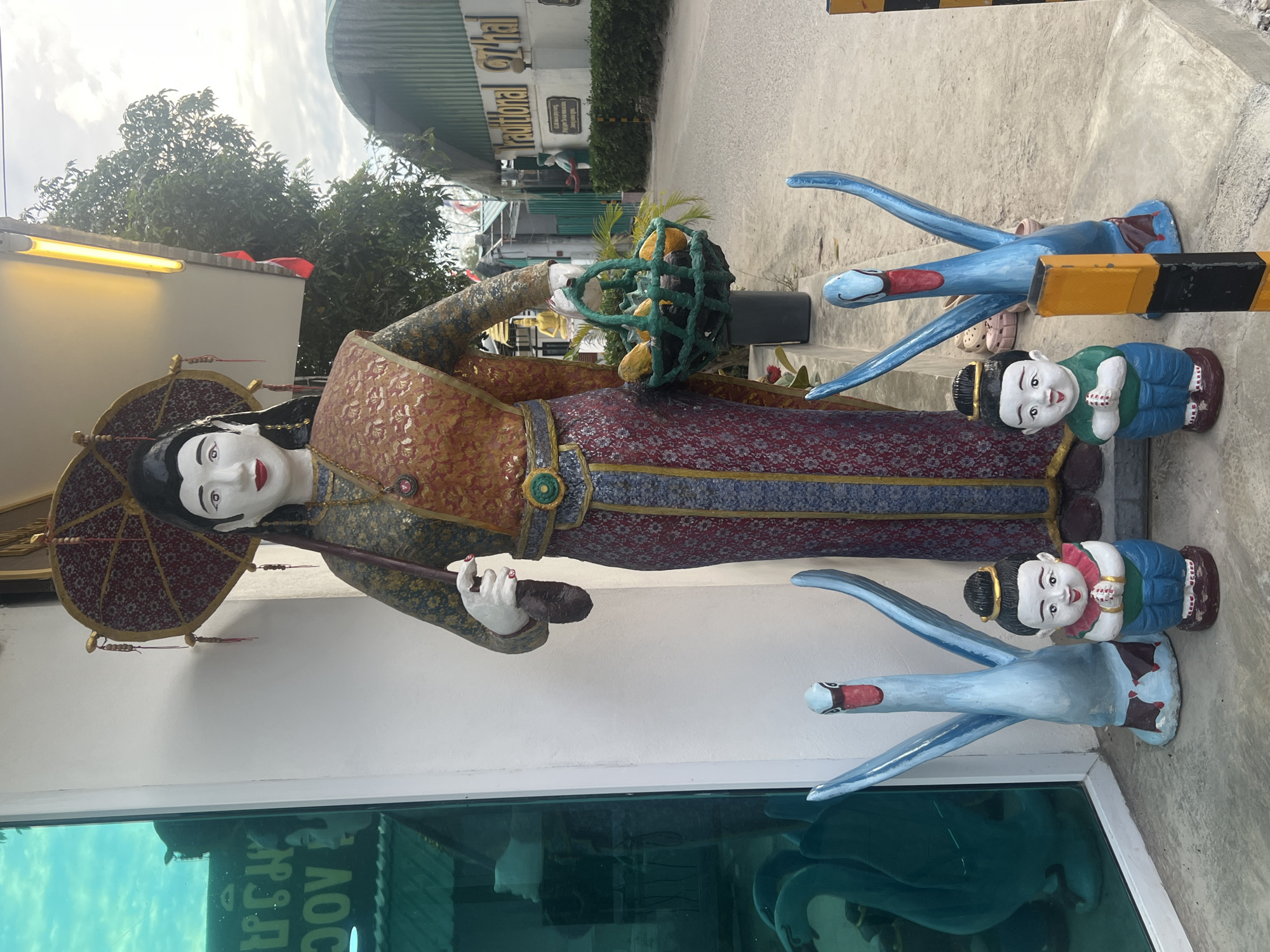 This is a full-shot, eye-level, outdoor photograph of several decorative statues and a building facade in the Ko Pha-ngan District, Thailand. The central subject is a large, life-sized statue of a woman dressed in traditional attire. She has dark hair pulled back and wears a red and gold patterned sarong, a gold-colored top, and a patterned sash. She holds a dark umbrella with a red and gold patterned canopy above her head with her left hand, and a green woven basket filled with yellow fruit in her right hand.
In front of the woman statue, slightly to her left, is a blue statue of a swan with its wings spread. To the woman's right, also slightly in front, is another blue swan statue. Between the two swan statues and in front of the woman are two smaller statues of children. Both children are depicted with hands pressed together in a prayer-like gesture. The child on the left wears a green top and blue pants, and the child on the right wears a red top and blue pants. The statues are positioned on a concrete surface.
To the left of the woman statue is a large, reflective turquoise-colored glass window. Inside the window, several blurry, dark blue duck or swan-like objects are visible.
Behind the statues and to the right, a building with a white facade is visible. A sign on the building reads "Traditional Thai". The building has a green, corrugated roof. Further in the background, trees and other structures line a paved road. The sky is cloudy.