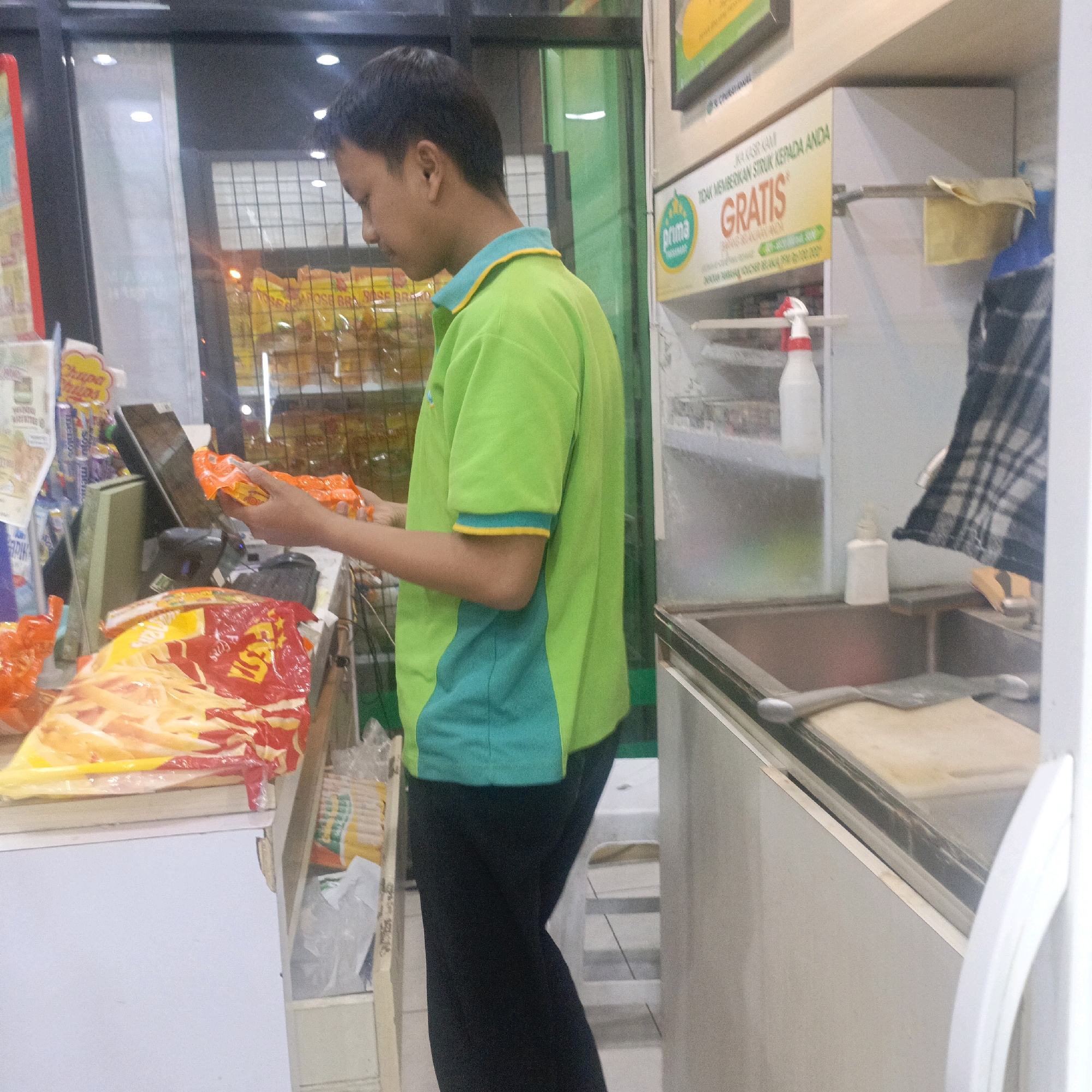 The image depicts a young male, likely an employee, standing behind a counter in what appears to be a convenience store or minimarket in Salatiga, Indonesia. He is wearing a bright green polo shirt with turquoise and yellow accents and dark pants. His attention is focused downwards as he handles two orange-colored snack packages.
In the foreground, a large red and yellow bag of snacks, possibly potato chips, is prominently displayed on the counter. To the left of the snack bag, a shelf holds various packaged goods, including what look like more snacks and other retail items. Behind the counter, a cash register or point-of-sale system is visible, along with shelves stocked with more merchandise, some of which appear to be food items in yellow packaging.
To the right of the boy, a promotional sign with the text "GRATIS" (meaning "Free" in Indonesian) and a logo for "Prima" is displayed on the wall. Further to the right, a sink area is visible, with a bottle of cleaning spray and a dishtowel hanging nearby. The overall environment suggests a functional retail space with displays of products and operational elements like a sink and point-of-sale equipment. The lighting suggests it is daytime, likely indoors.