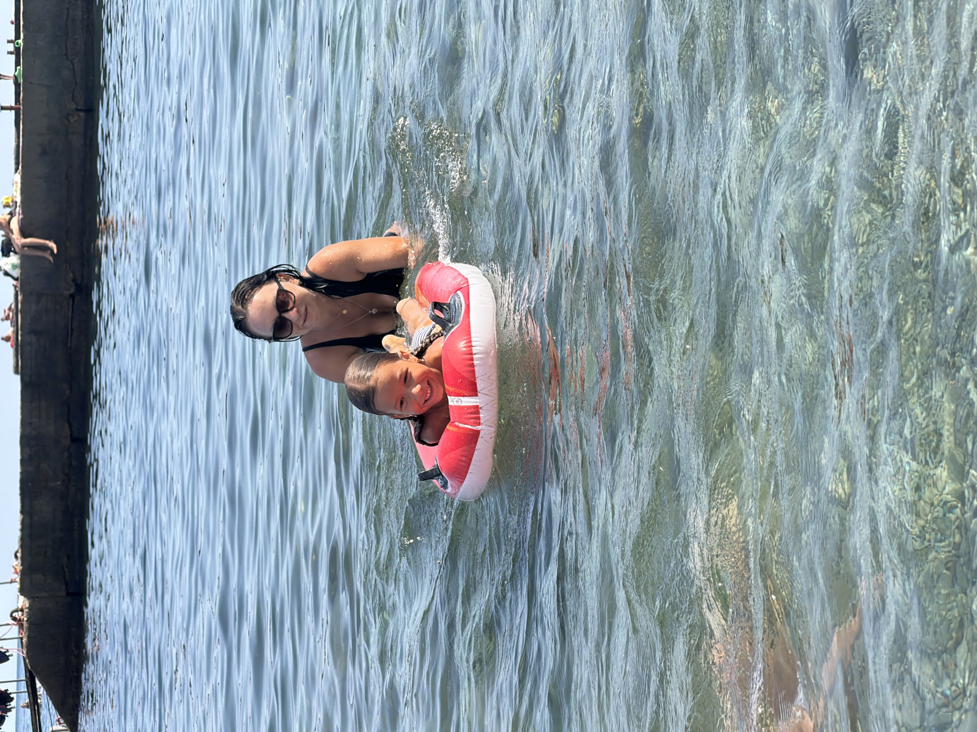 In the city of Partenit, Ukraine, a woman and a young girl are in the sea. The girl is floating on a red and white inflatable ring, smiling. The woman, wearing a black swimsuit and sunglasses, is behind her, in the water. In the background, a concrete pier juts out into the water, with several people visible on it. The water is clear and calm, with a visible seabed.