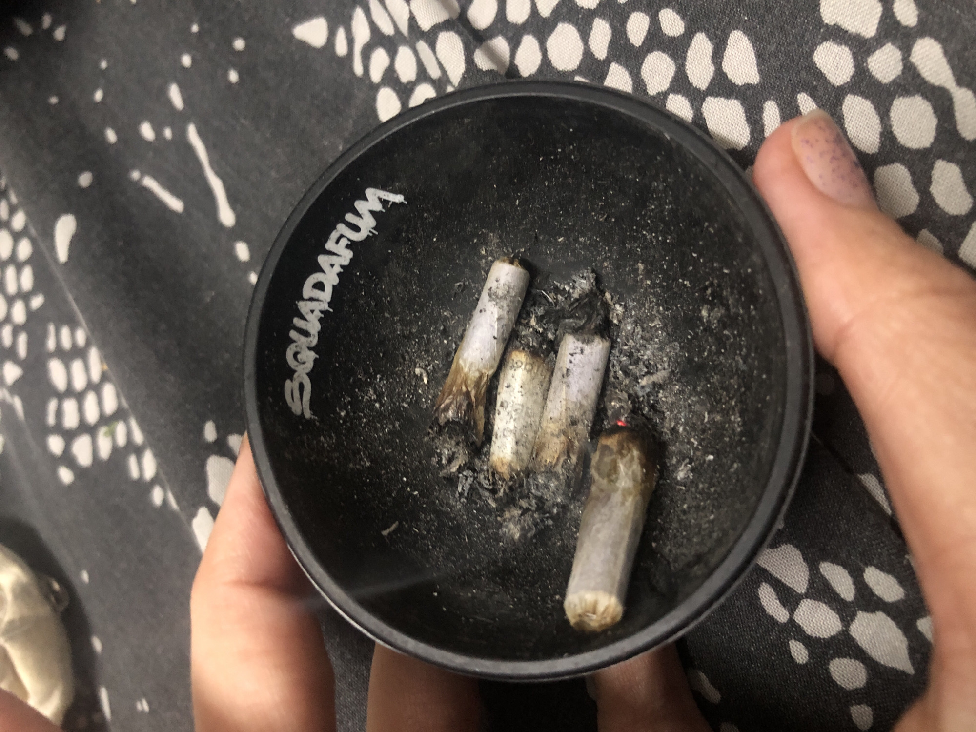 A close-up shot shows a black ashtray held in someone's hands. Inside the ashtray are three smoked marijuana joints. The ashtray has the word "Squadafum" written on the side. The background is a patterned fabric. There is no evidence to determine the location of the image, so the location requested cannot be ascertained.