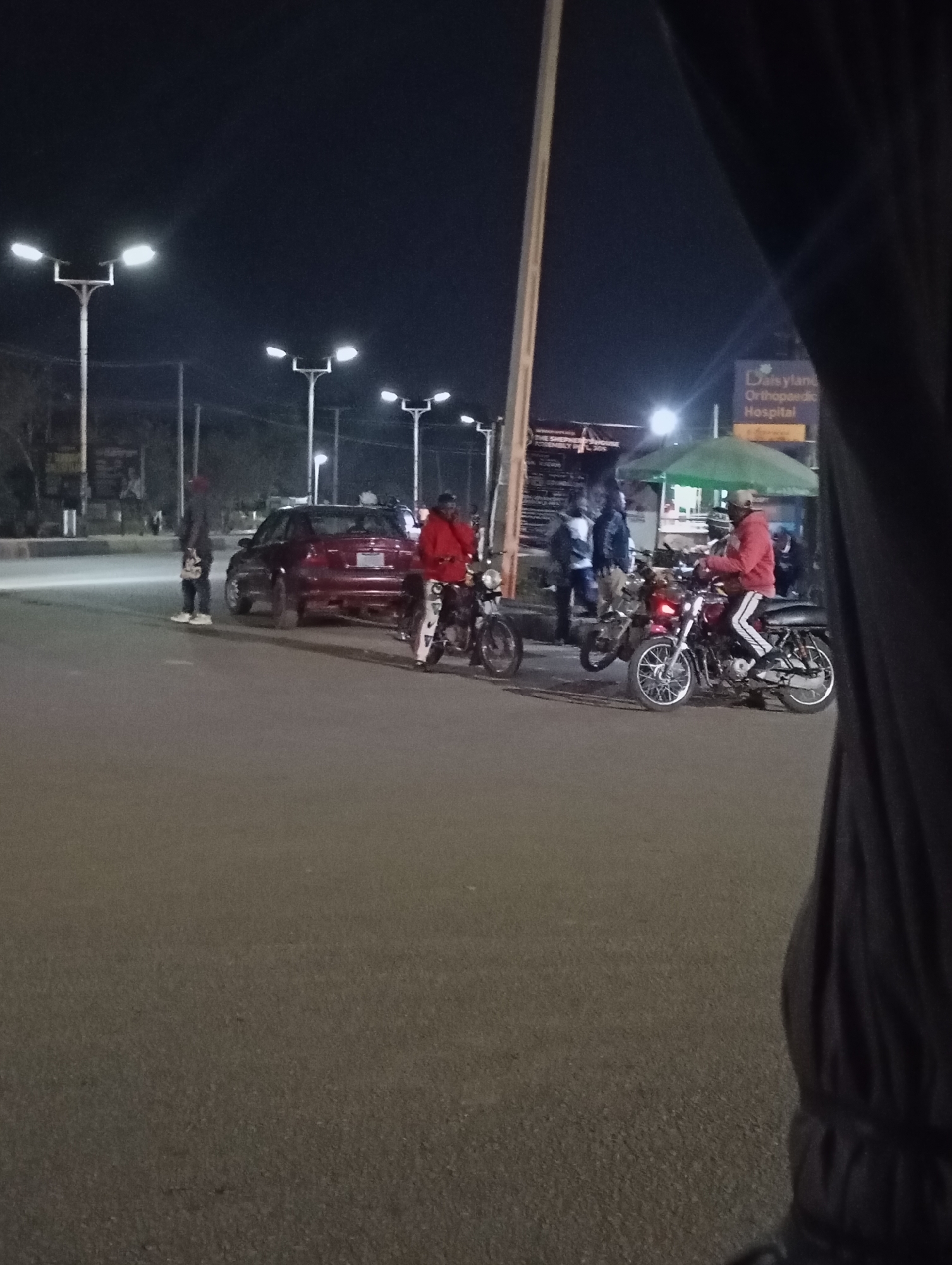 The image shows a street scene at night in Jos, Nigeria. The location is identifiable by the "Daisyland Orthopaedic Hospital" sign in the background.
Several people are present, including individuals standing near a red car and others near motorcycles. Two motorcyclists are visible on their bikes, wearing red tops and light colored pants with stripes. Streetlights illuminate the area. The road surface is asphalt. The scene appears to be an active street with several elements including vehicles and people congregating. A structure covered with a green canopy is visible behind the motorcycles. Part of a person is visible at the right side of the image, obscuring the scene.