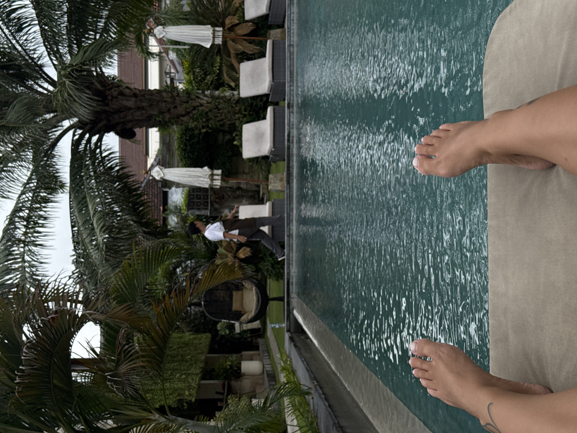 The image captures an outdoor scene at what appears to be a tropical resort in North Kuta, Indonesia, viewed from a low perspective. In the immediate foreground, two bare feet and lower legs rest on a light-colored lounge cushion, indicating a person relaxing by the pool. A tattoo is visible on the ankle of the left foot.
The central feature is a swimming pool with dark green, rippled water, suggesting an infinity edge or a dark bottom. The pool extends horizontally across the frame, with sunlight or diffused light reflecting off its surface.
Beyond the pool, a paved path or lawn leads into a lush, green landscape dominated by several large palm trees with dense fronds. Amidst this greenery, a person dressed in a white shirt and dark trousers is walking away from the viewer, towards the left side of the frame. Nearby, a distinctive round, wicker-style hanging chair with a light cushion adds to the relaxed ambiance.
The background reveals more resort amenities, including several light-colored lounge chairs with white cushions, positioned near the pool and shaded by white parasols. Buildings are partially visible through the foliage, with an overcast or diffused sky overhead, suggesting a cloudy day or late afternoon. The overall setting evokes a serene and leisurely vacation environment. There is no visible text in the image.