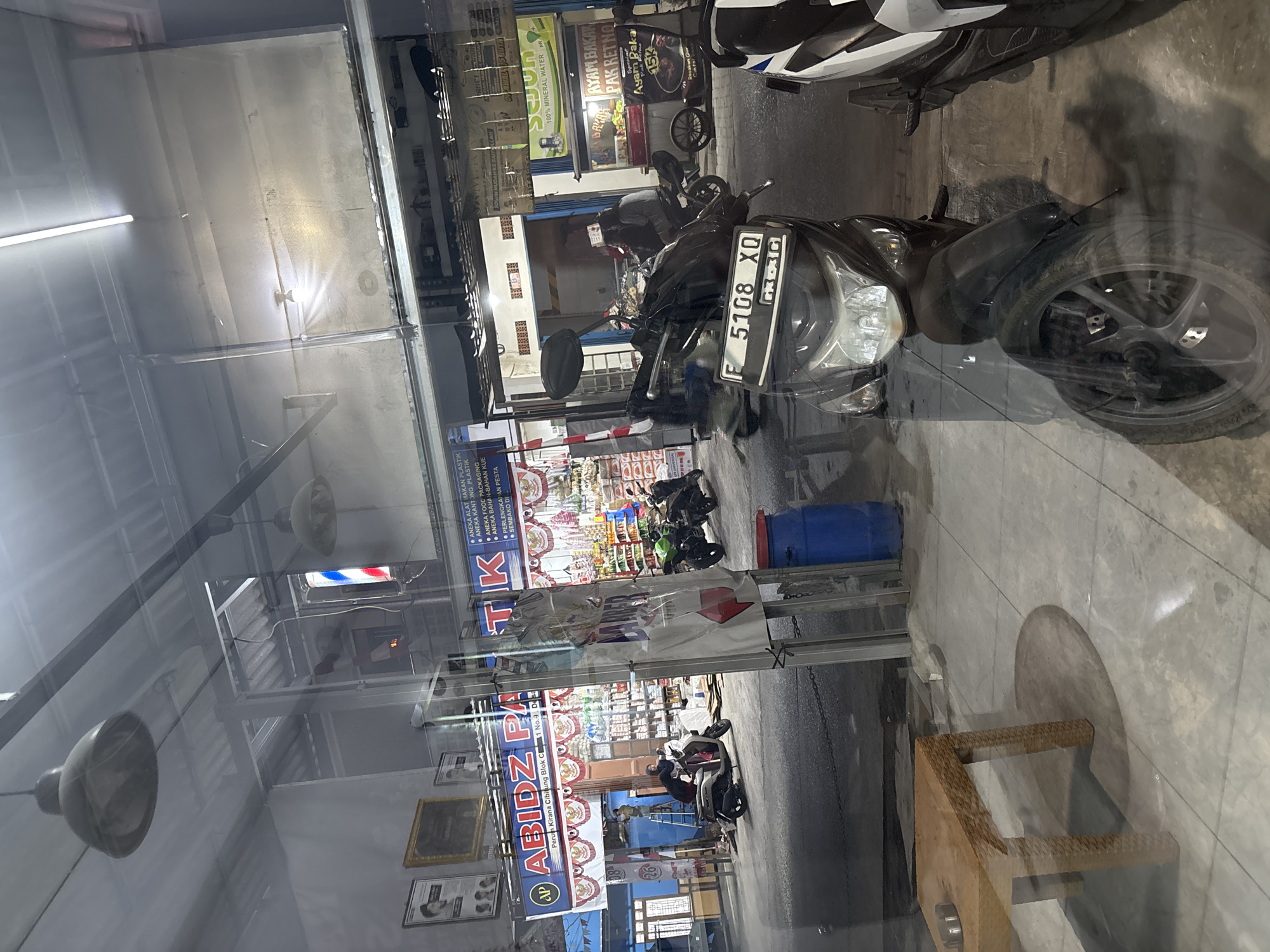 This image captures a street scene in Wanajaya, Indonesia, as viewed through a reflective surface, likely a window. The foreground is dominated by the reflection of a dark-colored scooter, its license plate clearly visible as "F 5108 XQ". To the right, a portion of another scooter, predominantly white with black accents, is also visible.
The mid-ground reveals a bustling street scene. Several motorcycles are parked or in motion. One scooter, a lighter color, has a person wearing a headscarf riding it. In the background, a shop with a large banner reading "ABIDZ PATIK" is prominent, featuring red and white decorative elements. Adjacent to this shop is another establishment with shelves stocked with various goods and a sign for "AYAM BAKAR PAK RETNO" which appears to be a food stall. A blue barrel is situated on the sidewalk near the street.
The environment suggests a commercial area, with shopfronts lining the street and a paved roadway. The lighting indicates it is likely late afternoon or early evening, with artificial lights from the shops illuminating the scene. The reflection in the foreground adds a layer of depth and visual complexity, showing elements of an indoor space, including a wooden table with an ashtray. The overall atmosphere is one of everyday urban life in Indonesia.