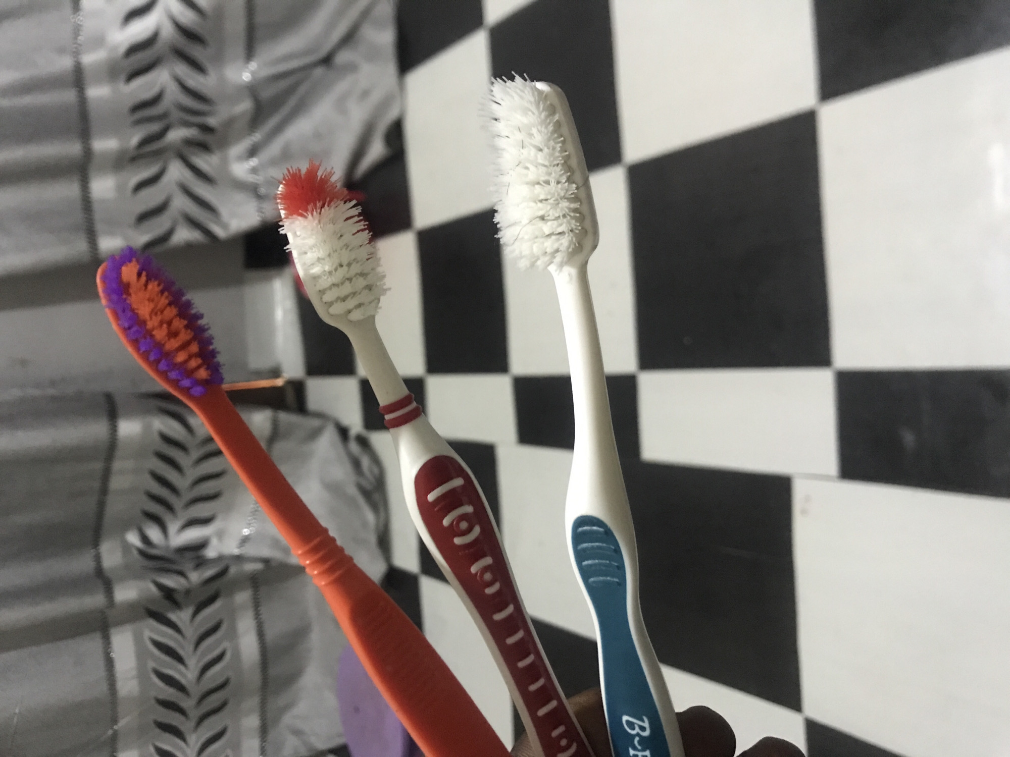 Three toothbrushes are displayed on a black and white checkered floor. The topmost toothbrush is orange with purple bristles. Below it is a toothbrush with a white handle and red bristles. The bottom toothbrush has a white handle with blue accents. A patterned curtain is visible in the background.