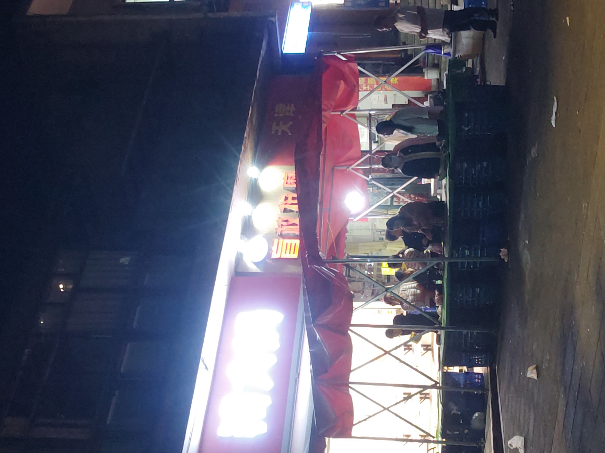This media file captures a nighttime street scene in what appears to be a busy commercial area, likely in Chongqing High-tech Zone, China, given the context. The image is oriented vertically (rotated 90 degrees clockwise).
On the left side, a brightly lit storefront dominates, featuring multiple luminous signs. The most prominent sign, positioned high, is a long, blue-white rectangular light. Below it, a red banner or awning displays Chinese characters, with "天堂" (Tiāntáng, meaning "Heaven" or "Paradise") clearly visible. Further down, another large, overexposed red and white sign emits bright light, making its text unreadable. Three round, white lights are also affixed to the building facade.
To the right, an informal gathering of people is visible under a red canopy supported by metallic scaffolding. Several individuals, mostly men, are present. Some are standing, while others appear to be seated on or around green and blue plastic crates, which might be serving as makeshift stools or tables. They are engaged in various activities, possibly eating, conversing, or waiting. Their faces are largely obscured or blurred due to the low light conditions and distance. One person in a light-colored shirt and dark trousers stands near the far right, overlooking the scene.
The ground is a dark, paved surface, with some scattered debris or trash visible. The overall setting suggests a bustling evening, possibly at a street food stall or an outdoor extension of a shop, characterized by casual interaction and a vibrant, illuminated atmosphere against the dark night sky.