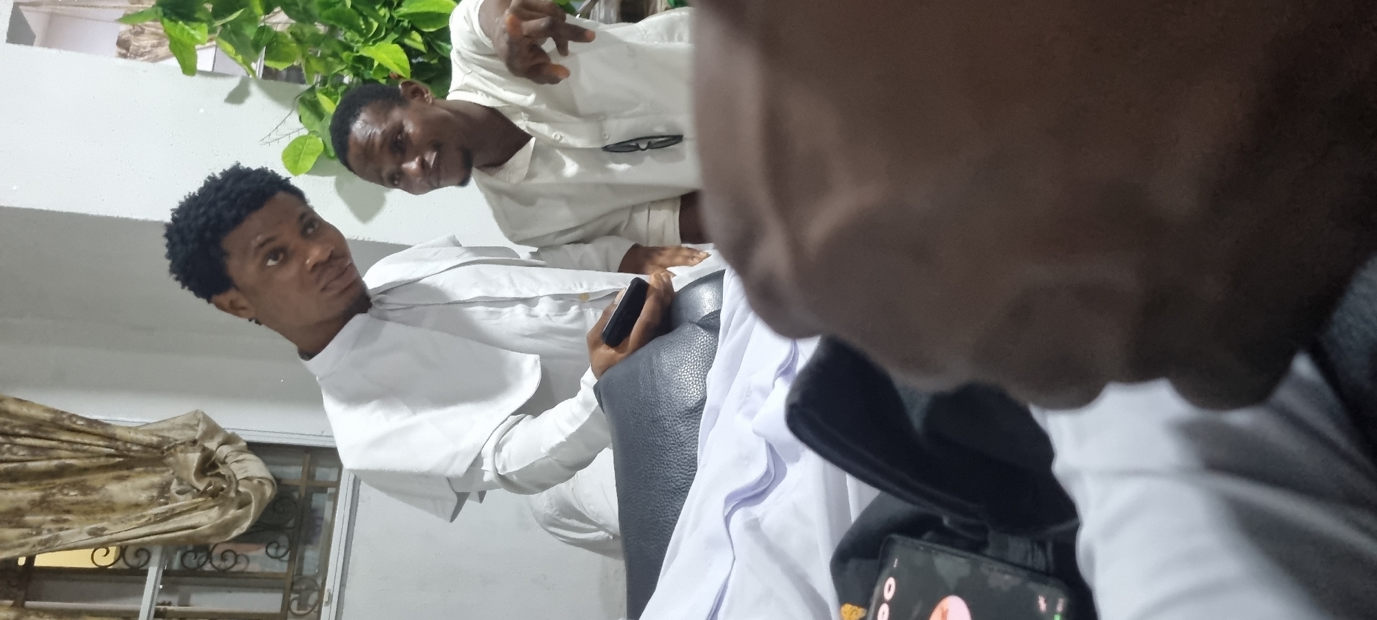 Two young men, likely in their late teens or early twenties, are present in an indoor setting. The man on the left is wearing a white garment with a cape-like overlay, possibly a uniform or religious attire. He is holding a black mobile phone in his right hand. The man on the right is wearing a light-colored button-down shirt. He is gesturing with his right hand, appearing to point. A pair of dark-rimmed glasses is attached to the front of his shirt.
The background features a window with draped curtains on the left and a potted plant with green leaves on the right. The overall environment appears to be a room, possibly a living space or communal area. A dark-colored bag or piece of furniture is visible in the foreground. The perspective suggests the photo was taken from a low angle, with the photographer's hand or arm partially obscuring the bottom portion of the image. The lighting is moderate, creating a slightly dim atmosphere. There are no discernible urban or city-specific features in the image.