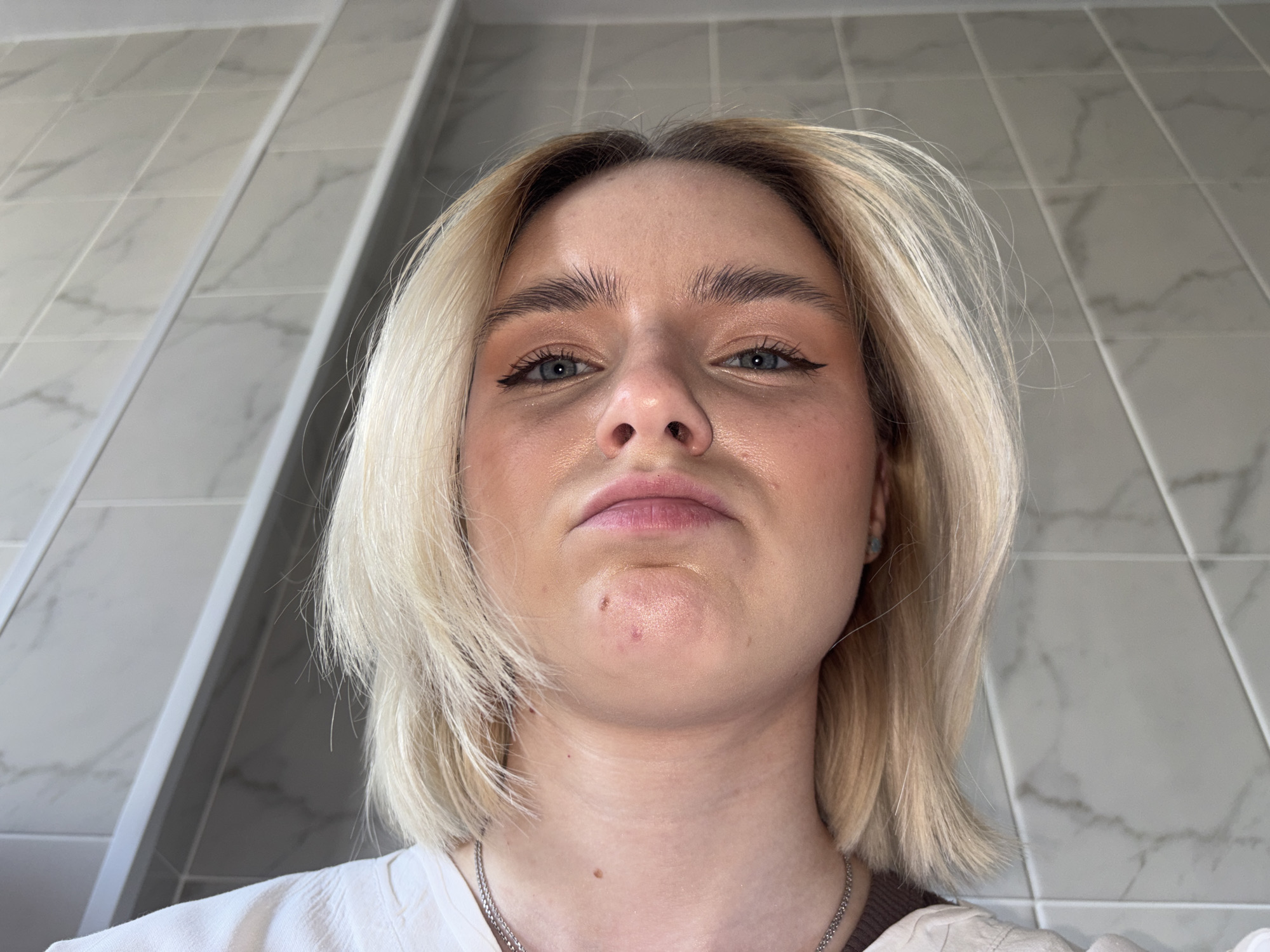 An upward-angled selfie captures a young woman looking directly into the camera from below. She has chin-length blonde hair with visible darker roots and light-colored eyes. Her expression is neutral, with closed lips and a slightly raised chin. She wears makeup, including black winged eyeliner and light eyeshadow, and her natural skin texture is visible, showing some shine on her forehead and nose, along with a few small red marks on her chin. Around her neck is a silver-colored chain necklace, and she is wearing a light-colored top, possibly white, with a hint of a darker, perhaps beige, garment on her right shoulder. The background consists of light grey or white rectangular tiles featuring a subtle marble-like pattern, arranged in a grid on a wall. This tiled setting suggests an indoor location. There is no visible text in the image. The provided context for the location is Saint Petersburg, Russia.