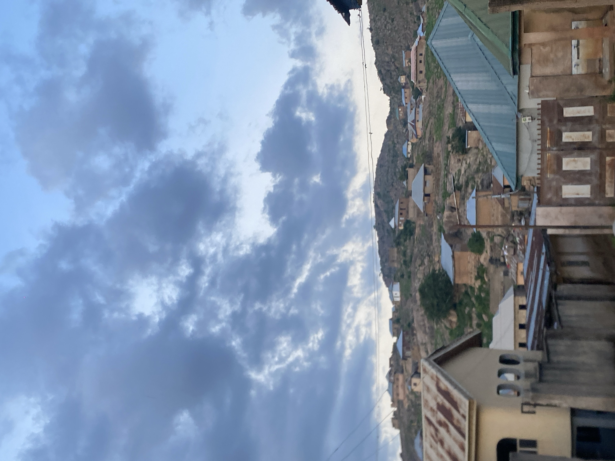An image, oriented vertically, captures a panoramic view of a cloudy sky above a settlement nestled on a hilly, rocky landscape, likely in Jos, Nigeria.
The upper portion of the frame is dominated by a vast expanse of light blue sky, largely covered by a dramatic array of grey and white clouds of varying densities. Some areas of the sky show brighter patches, suggesting diffused sunlight either breaking through or illuminating the clouds from a low angle.
Below the sky, the rugged terrain of hills or mountains is visible, characterized by earthy tones and sparse vegetation. Numerous houses and structures are built into the hillsides and across the slopes. These buildings appear to be primarily one or two stories high, constructed with materials like concrete or mud brick, and many feature distinctive corrugated metal roofs, some painted green, others showing signs of rust. The houses are densely packed in some areas, creating a mosaic across the landscape.
Thin power lines or utility wires traverse the middle ground of the image, stretching horizontally against the sky and hills, indicating infrastructure within the community. In the foreground, parts of several buildings are more clearly visible, including one with a corrugated metal roof and another with a beige facade and recessed window openings.
The soft, diffused lighting suggests the time of day is either early morning or late afternoon, possibly twilight, with the sun low or obscured by clouds. The weather appears overcast but calm. No specific human activity is discernible, and there is no visible text within the frame. The overall scene depicts a resilient settlement integrated into a challenging yet picturesque natural environment.