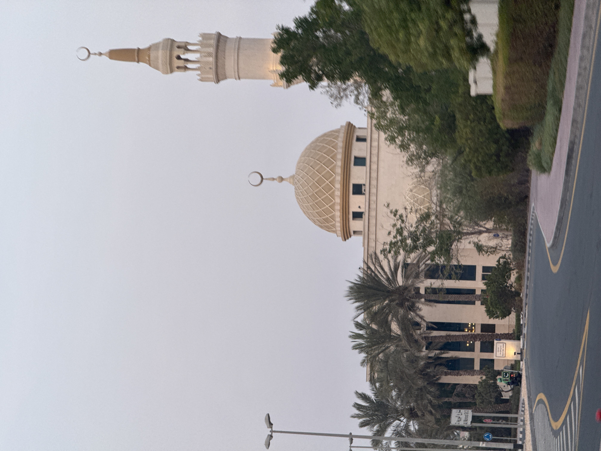 A mosque with a dome and minaret is visible on a cloudy day in Dubai, United Arab Emirates. The building is light-colored with a golden dome adorned with a crescent moon. The minaret, also light-colored, has several levels and is topped with a crescent moon. The mosque is surrounded by palm trees and other greenery. In the foreground, there is a road with yellow lane markings and a pedestrian walkway. A street sign with Arabic script and "Al Worood St 2" is visible on the left side of the image. A small green delivery vehicle is parked near the sign. The overall impression is one of a peaceful and well-maintained urban environment.