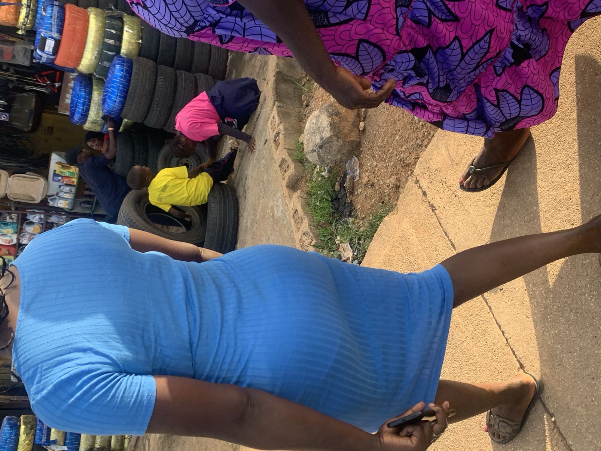 A street scene in Jos, Nigeria, features two women and three children in a dusty outdoor setting, with stacks of tires and shelves of goods in the background. In the foreground, a woman in a blue ribbed dress walks away from the camera, her back and left side visible. She holds a dark flip phone in her left hand. To her right, another woman is partially visible, wearing a vibrant pink and purple patterned dress.
Further into the scene, three children are interacting amidst stacks of tires. One child in a bright yellow t-shirt with "UNITY HOUSE" printed on it sits on a tire, looking towards another child. This second child, wearing a pink t-shirt and a dark skirt, is kneeling and appears to be examining something on the ground. A third child, partially obscured by tires and the kneeling child, is seen near a man wearing a dark shirt and a cap, who leans over a stack of tires, possibly interacting with the children.
The background reveals a store or market stall with shelves filled with various products, including bottles and containers. The ground is a mix of dirt, sand, and some patches of dry grass. The lighting suggests it is daytime, with some shadows cast by the objects and people.