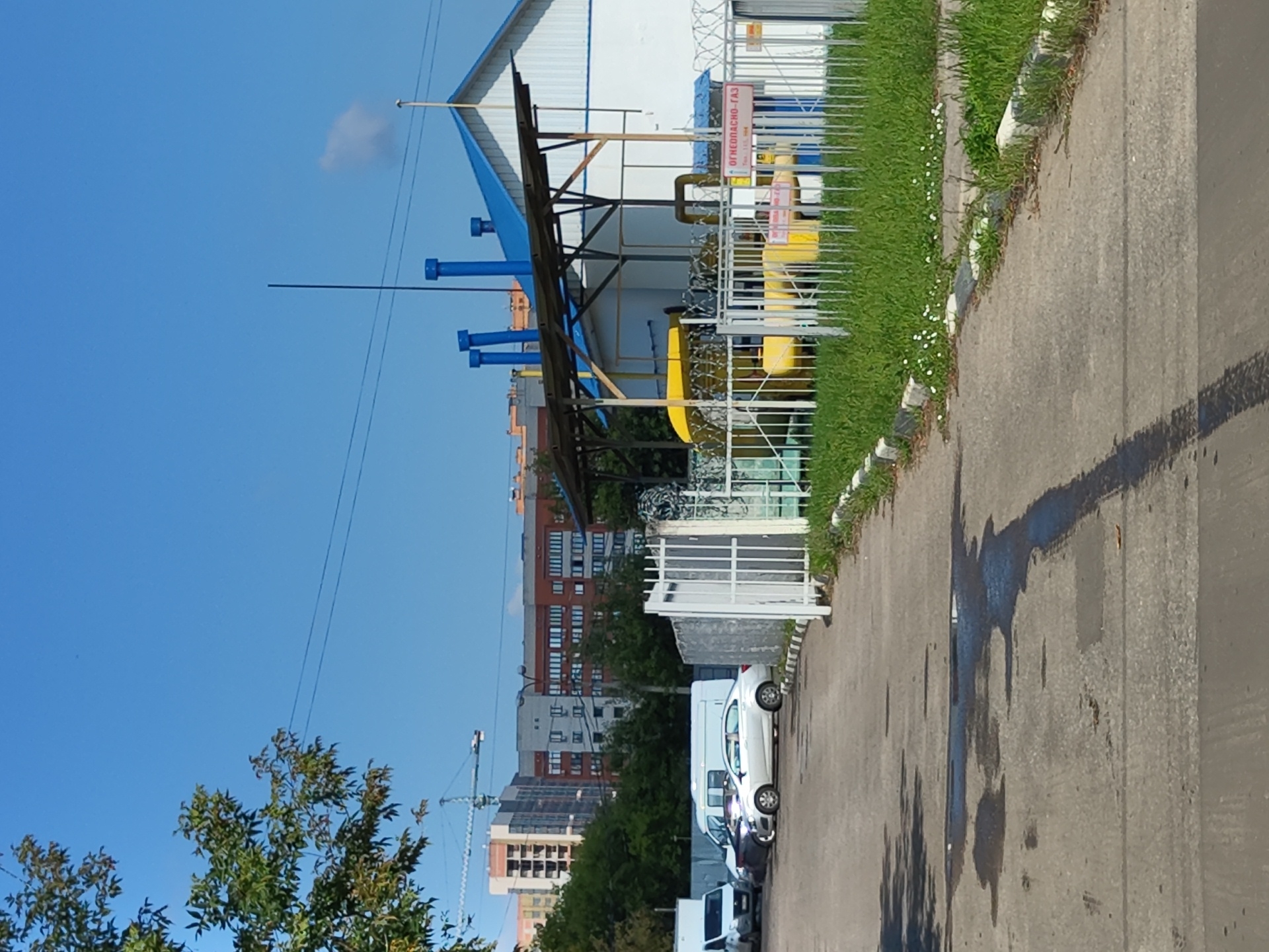 The image depicts a clear, sunny day in an industrial or utility area in Zhukovsky, Russia. In the foreground, a paved asphalt surface is visible with several puddles of water and tire marks. To the left, there is a white van and a silver sedan parked on the asphalt. A line of white metal fencing borders the scene on the right, with a strip of green grass and small white flowers along the base. Beyond the fence, a building with a white facade and a blue roof is situated, featuring blue vertical pipes and a yellow structure covered by a wooden-framed awning. A sign with red text reading "ОГНЕОПАСНО-ГАЗ" (Danger - Gas) is attached to the fence in front of this building. In the background, several multi-story residential buildings and a construction crane are visible against a bright blue sky.