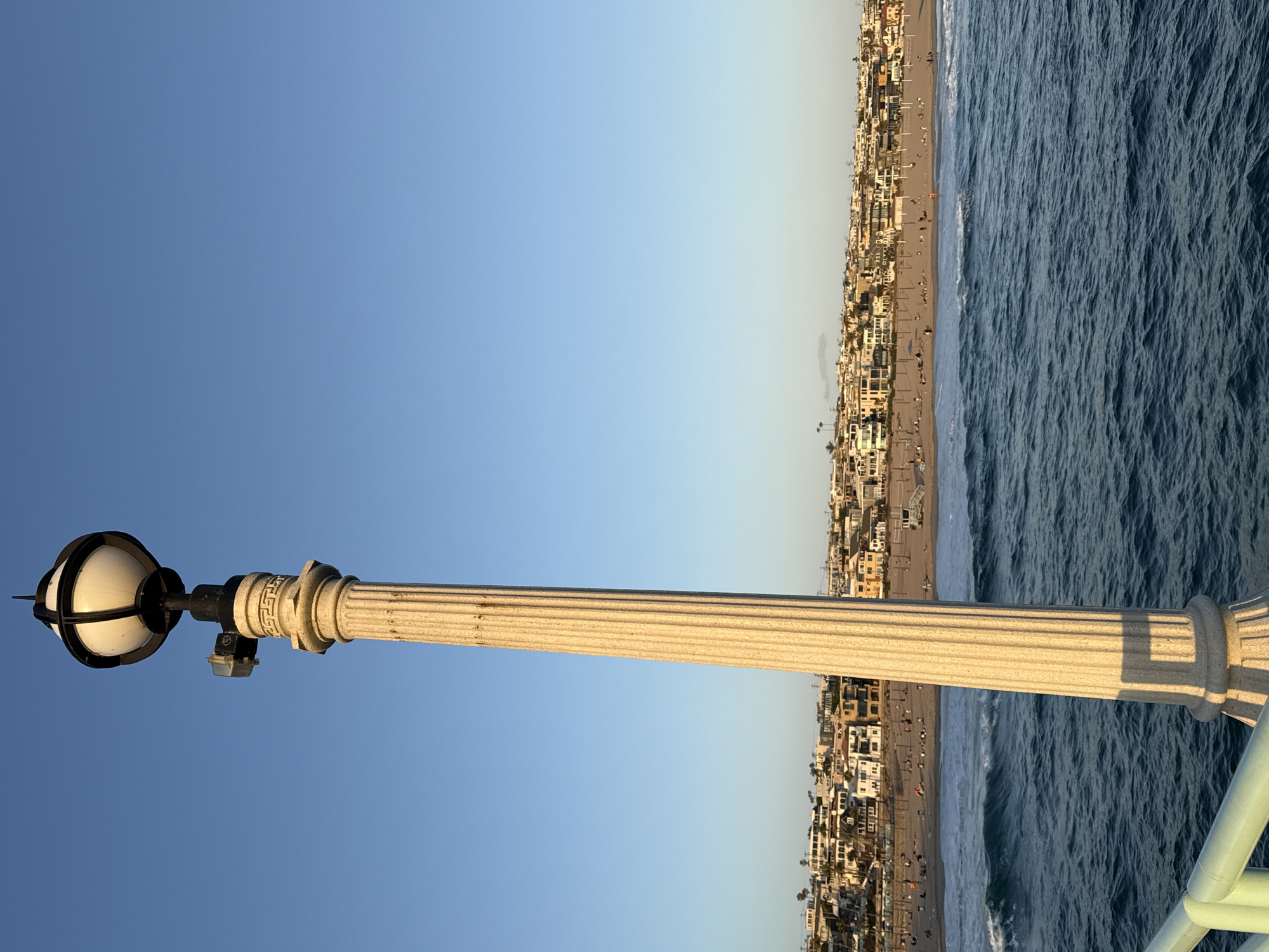 A lamppost stands prominently in the foreground of this vertical, medium-shot image. The lamppost is made of a light-colored, fluted material and is topped with a white, spherical light fixture encased in black metal. To the left of the light fixture, a small, square electrical box is attached to the lamppost. The lamppost casts a distinct shadow on the right side of its shaft.
The background reveals a wide, sandy beach populated by numerous people engaged in various activities, such as sunbathing and strolling. A lifeguard tower is visible near the center of the beach. The ocean in front of the beach exhibits moderate waves with whitecaps. Beyond the beach, a dense collection of buildings, primarily houses and multi-story residences, forms a backdrop that slopes upwards towards a clear, light-blue sky. The overall scene suggests a pleasant day at a coastal city. Based on the architecture of the buildings and the presence of palm trees, this location is likely Manhattan Beach, California, United States of America.