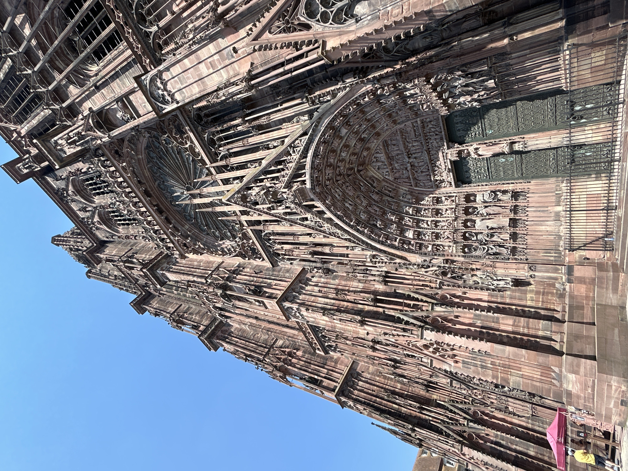 The image shows a low-angle, full shot of the Cathédrale Notre-Dame de Strasbourg in Strasbourg, France, under a clear blue sky. The cathedral's facade, constructed of reddish-pink sandstone, is highly ornate, featuring intricate Gothic architectural elements. These include numerous spires, statues, and a large rose window above the entrance. The main entrance is a large, arched doorway with double doors, surrounded by detailed sculptures and statuary. A black metal fence encloses the area in front of the doors. To the left of the cathedral, a person wearing a yellow shirt and white shorts stands near a red canopy tent, with another person partially visible behind a wooden structure. The overall impression is of the cathedral's imposing scale and detailed craftsmanship.